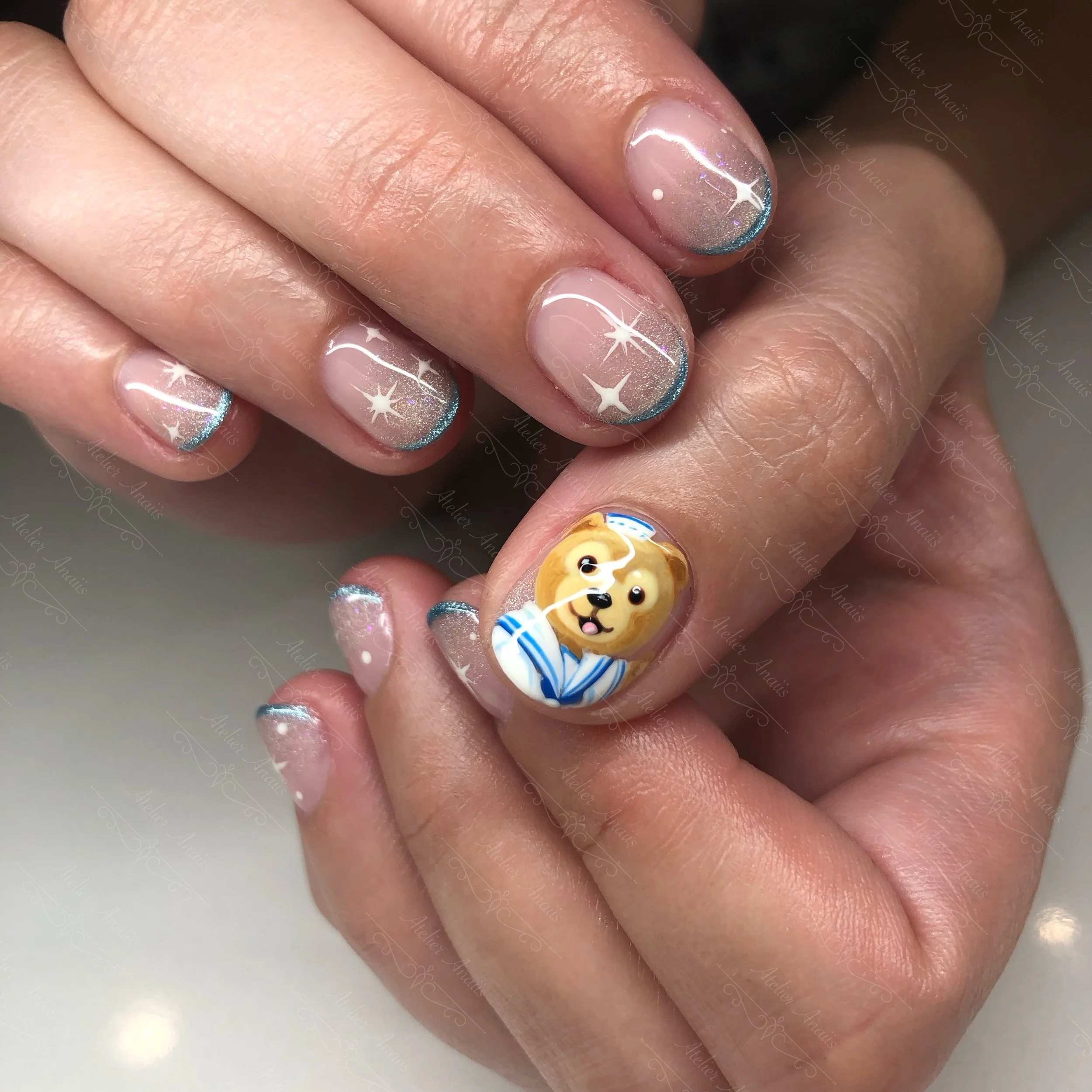 Hands with nails featuring intricate nail art with hand drawn teddy bear design and stars over a nude base with blue French tips.