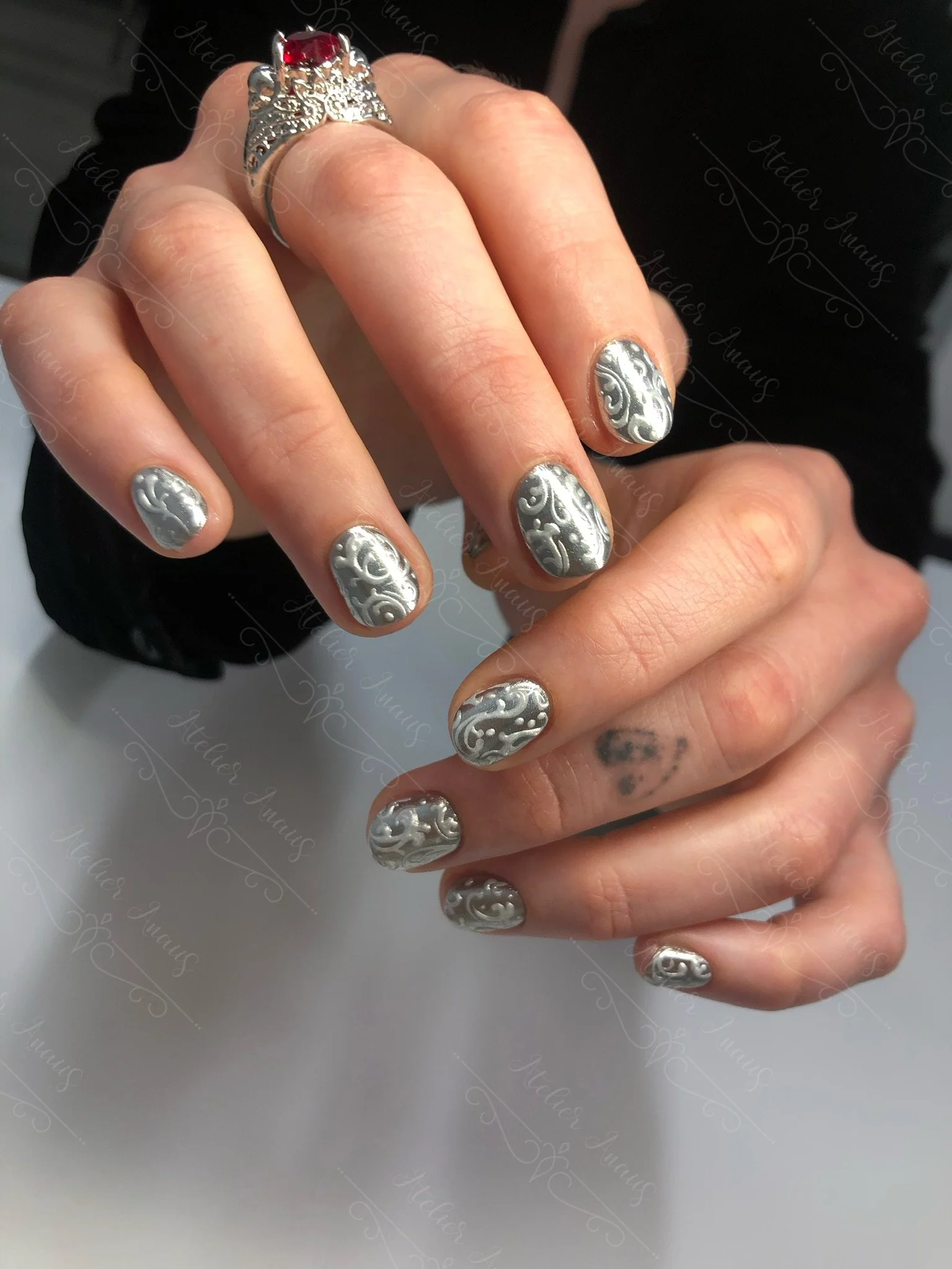 Hands with short nails featuring silver rococo style nail art embossed on muted silver base.