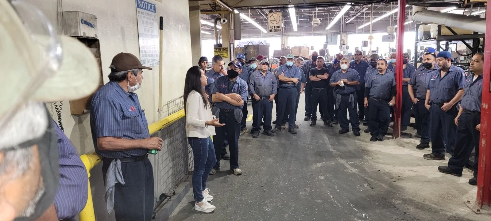 Grupo de empleados en un espacio de trabajo industrial, algunos con uniforme y mascarillas, escuchando a una mujer que está hablando.