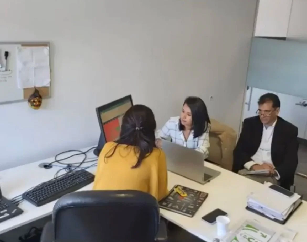 Tres personas en una reunión de oficina, dos mujeres y un hombre, sentadas en una mesa con laptops y papeles.