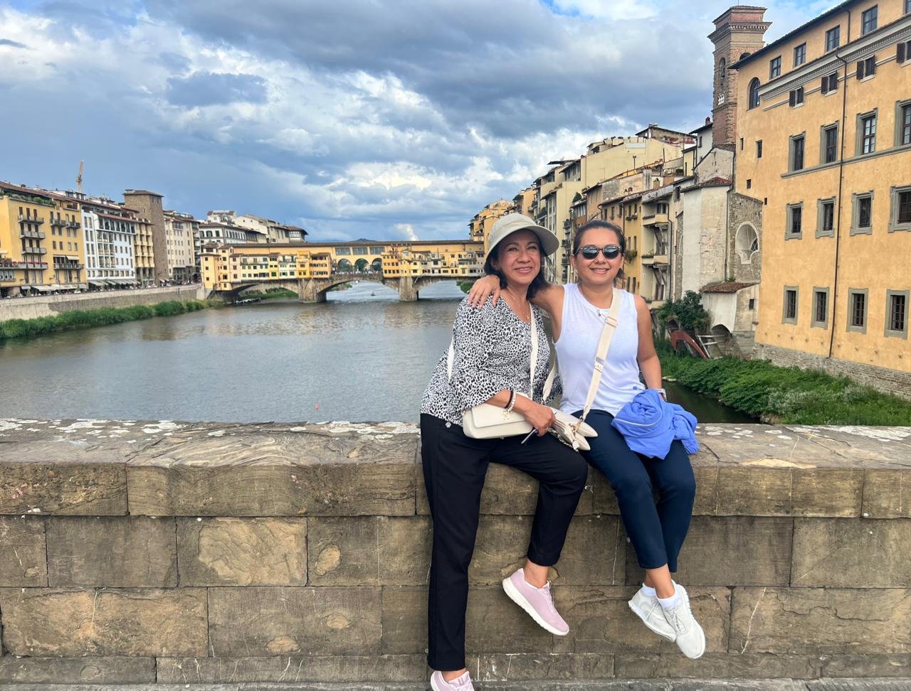 Dos mujeres sentadas en un muro de piedra junto a un río en Florencia, Italia, con edificios históricos amarillos y un puente en el fondo. Un cielo nublado complementa la escena con un ambiente pintoresco.