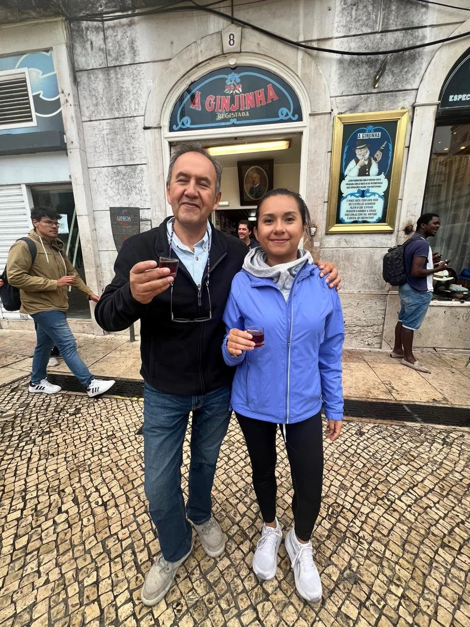 Dos personas, un hombre y una mujer, de pie en la calle frente a un establecimiento llamado 'A Ginjinha'. Ambos sostienen pequeños vasos con una bebida de color rojo y están sonriendo. La escena parece ser en un día nublado en una zona urbana.