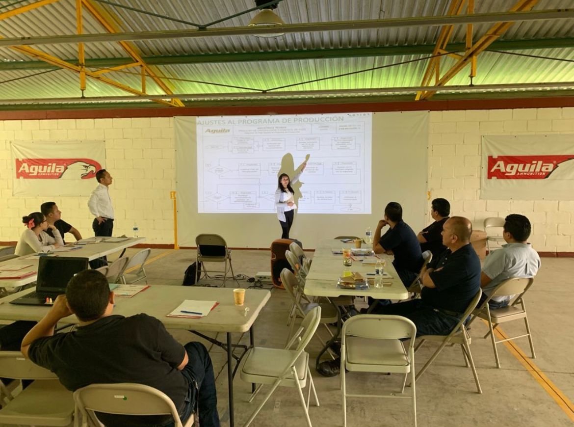 Presentación en una conferencia o reunión en un espacio cerrado, con personas sentadas en mesas observando a una mujer que explica en una proyección en la pared.