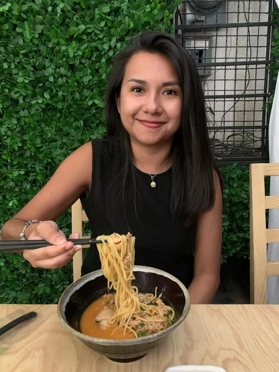 Mujer sonriendo con cabello oscuro sentado en una mesa, sosteniendo palillos y comiendo un tazón de ramen con carne y verduras, fondo con plantas verdes y pared con cajas eléctricas.