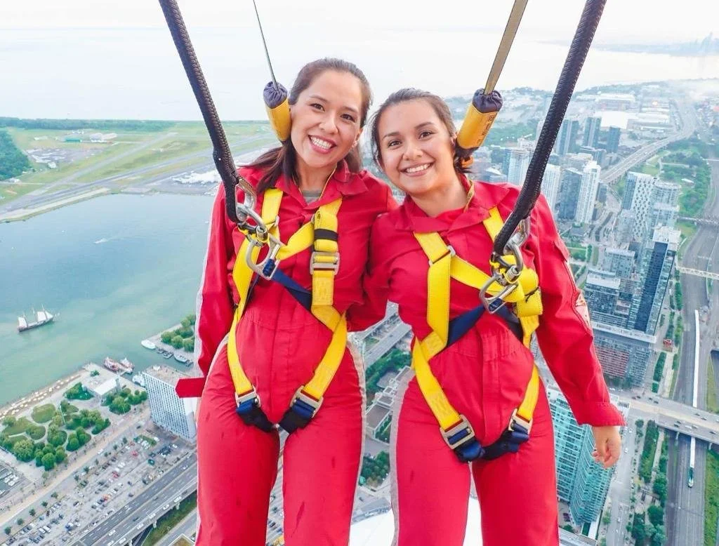 Dos mujeres sonrientes en un paracaídas, vestidas con trajes rojos y arnés, sobre la ciudad y un río.