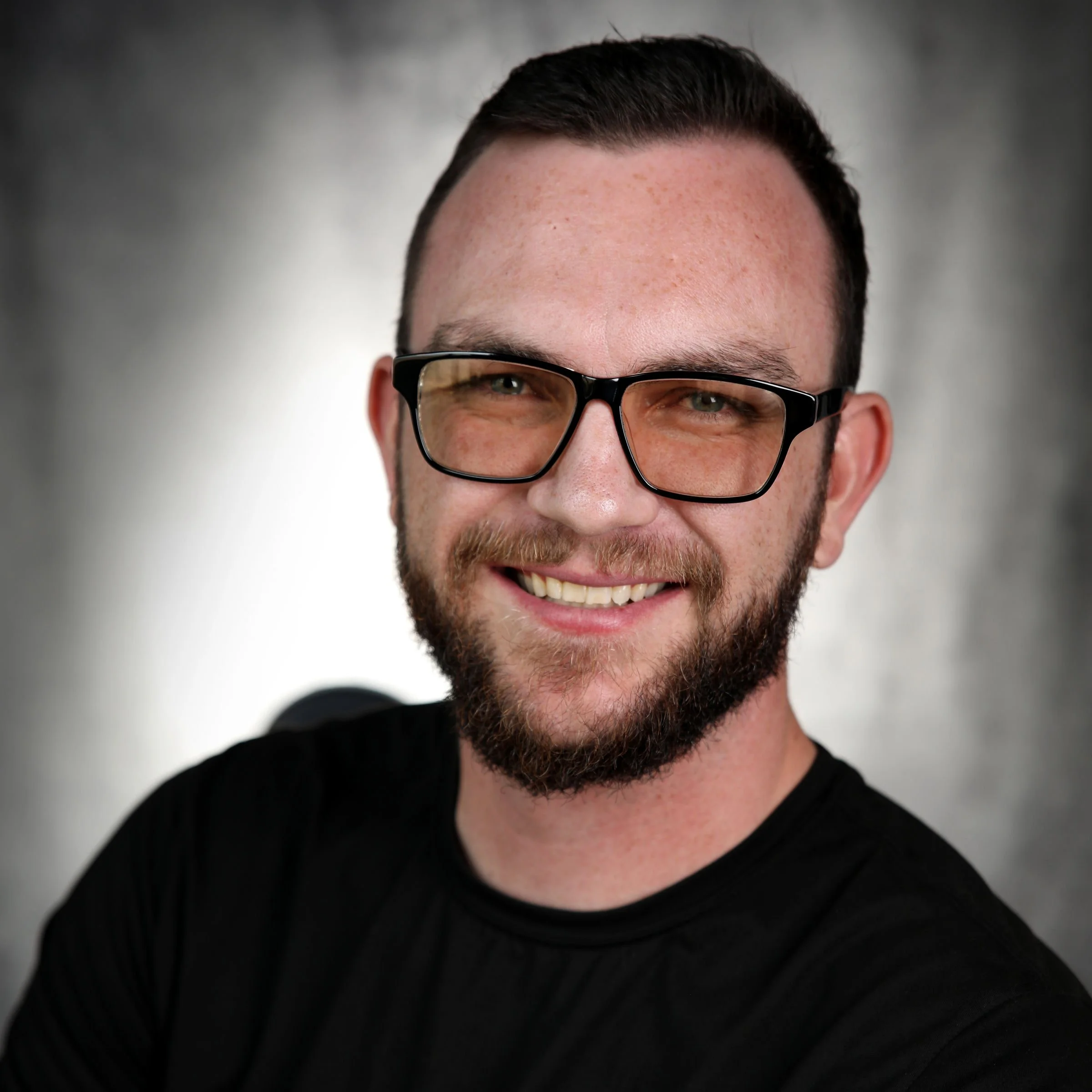 Close-up of a smiling man with glasses, beard, and short dark hair, wearing a black shirt, against a gray background.