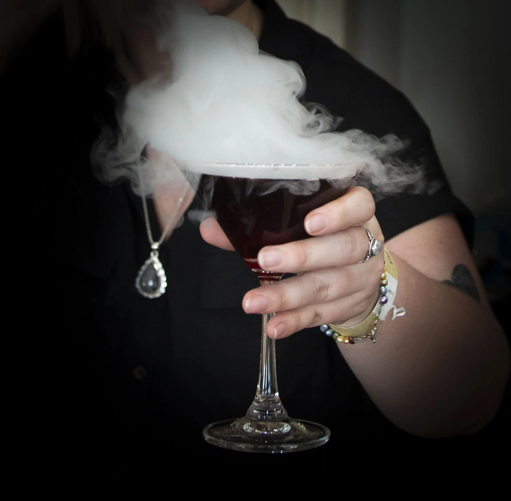 Close-up of a person holding a glass of dark beverage with smoke drifting over the surface of the liquid, wearing jewelry including rings and colorful bracelets.