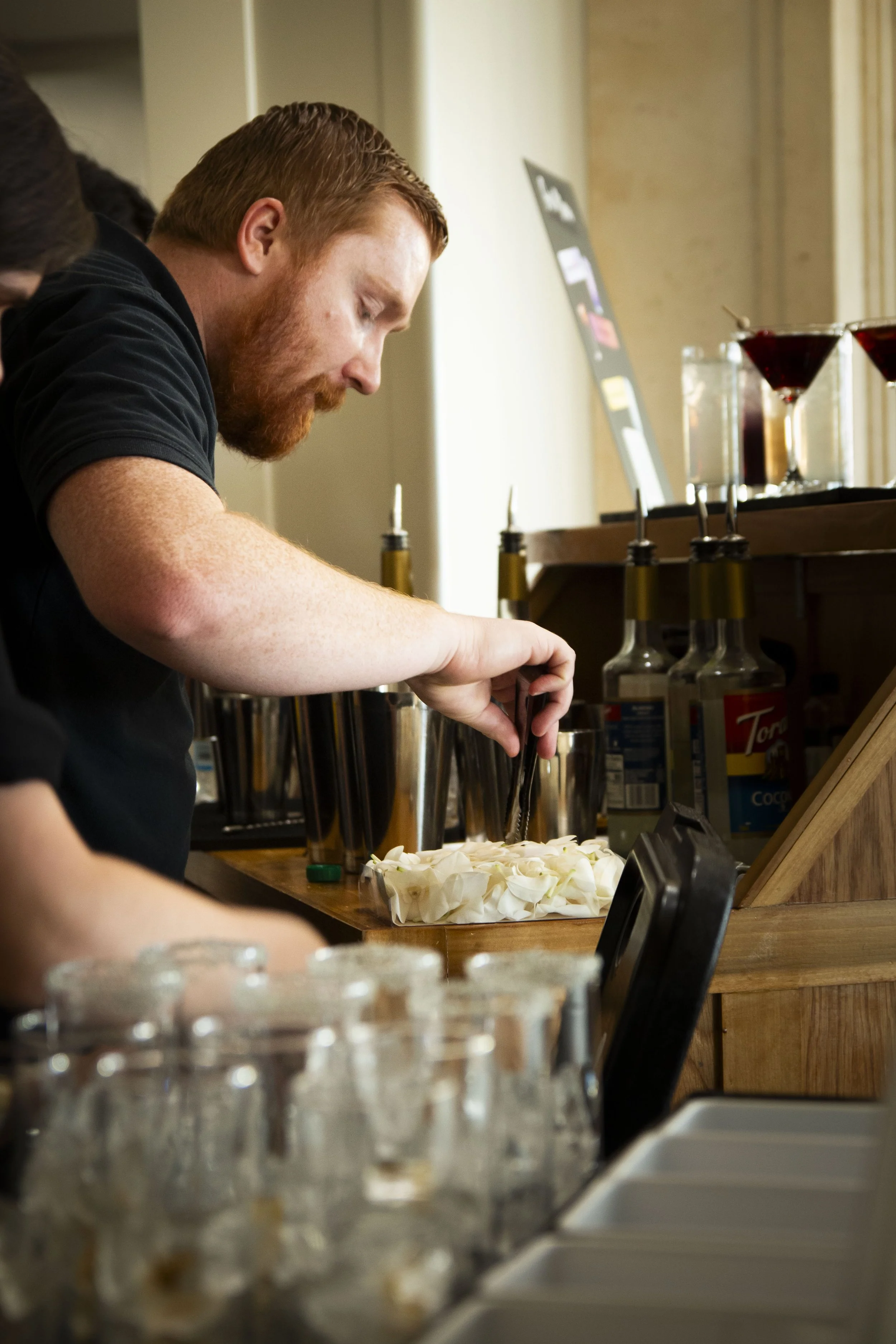 A man with red hair and a beard preparing mocktails at a classy bar, with glasses and bottles on the counter and a blurry row of glasses in the foreground.