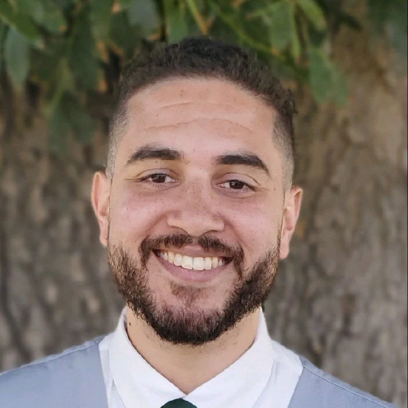 A smiling man with short dark hair, a beard, and mustache, wearing a light-colored shirt and a gray vest, standing outdoors with a tree and green leaves in the background.
