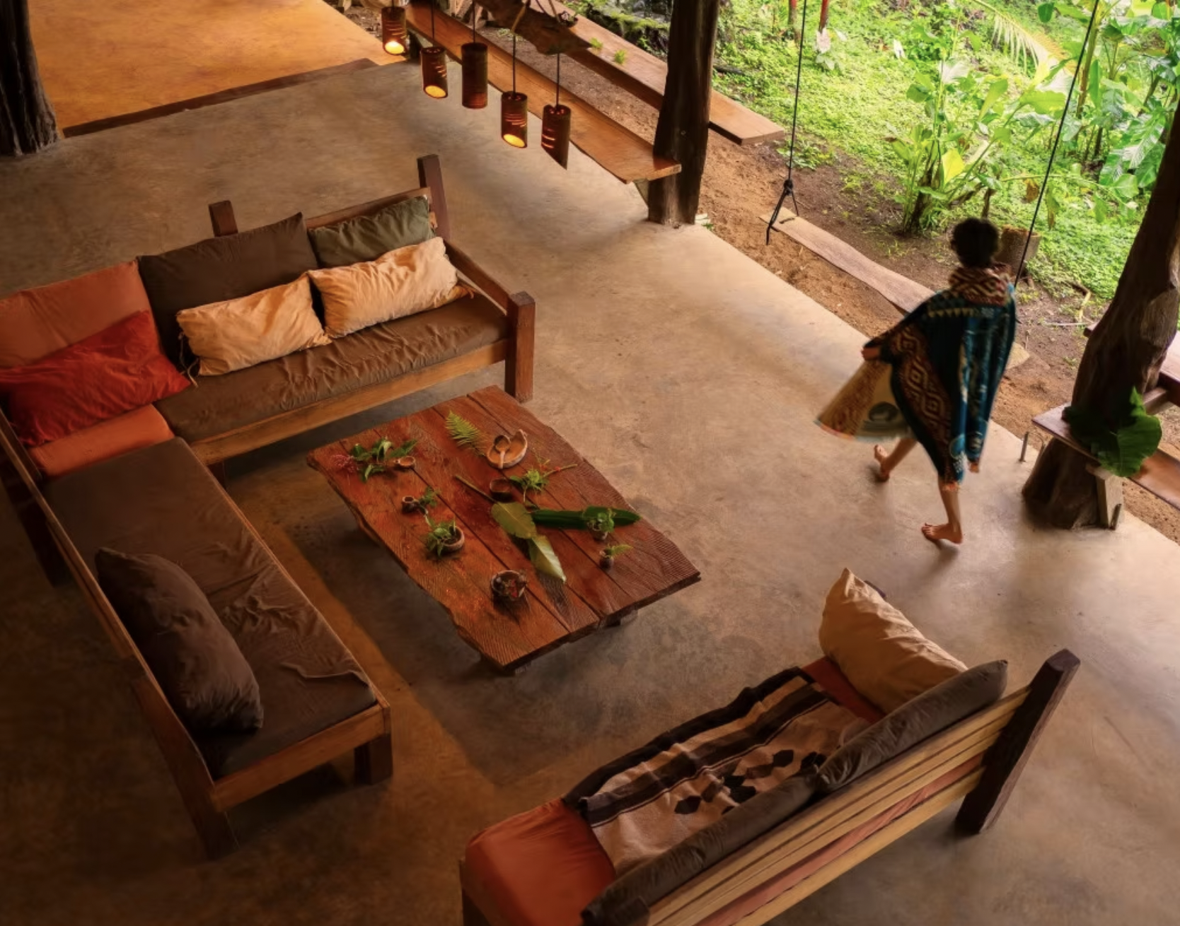 Living room with wooden furniture, cushions, and curtains, with a person walking along the outdoor patio area which has greenery and a swing.