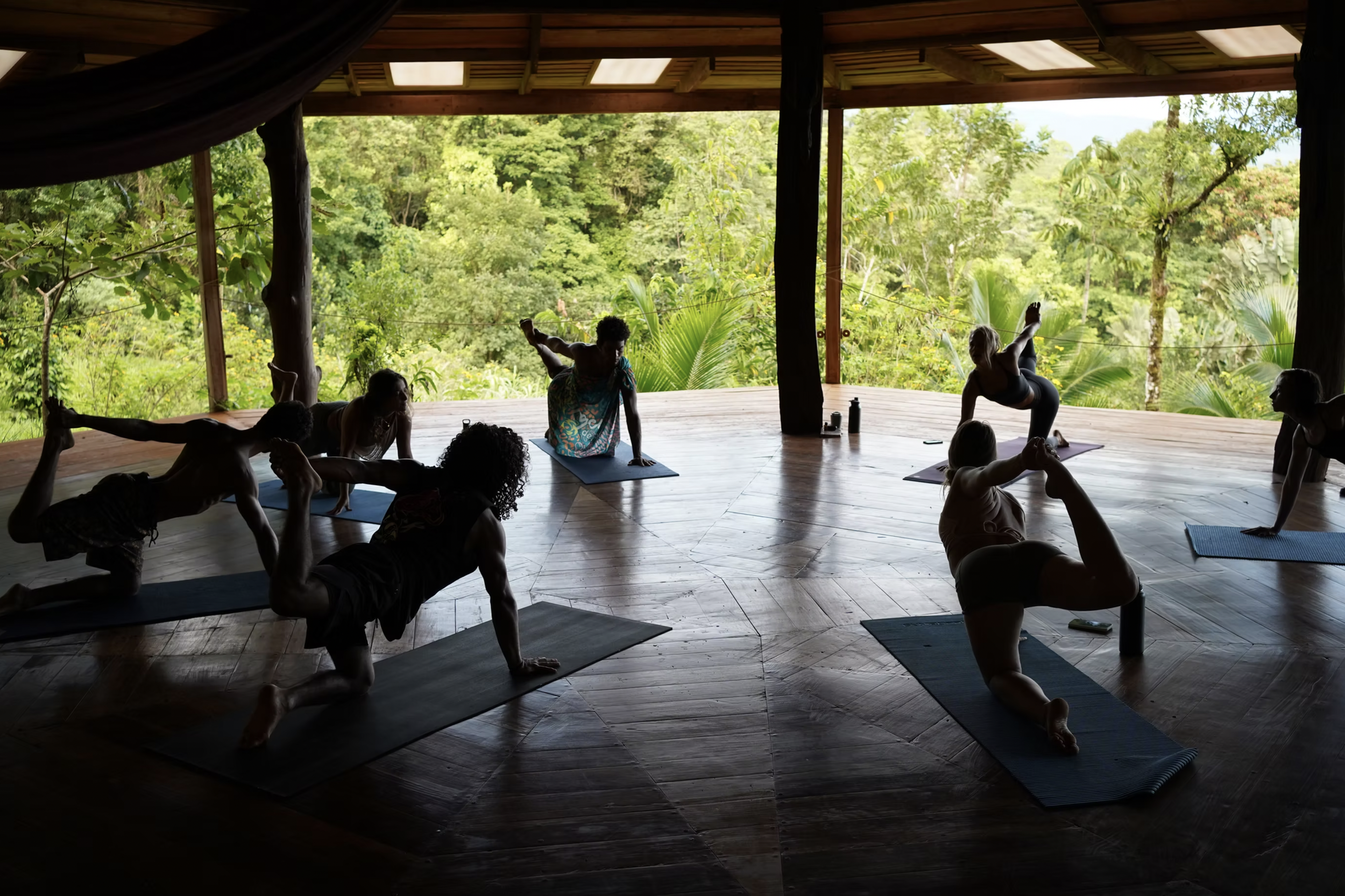 People practicing yoga on mats in a wooden, open-air room with a view of lush green trees.