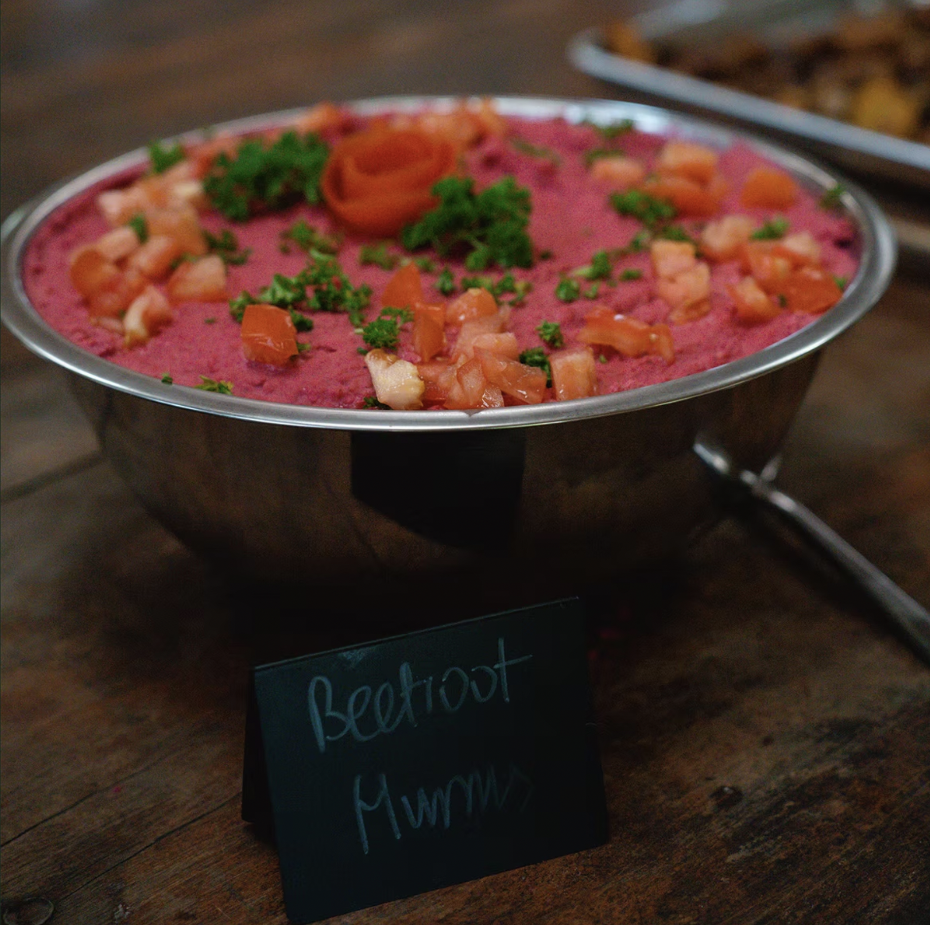 A metal bowl of beetroot hummus garnished with chopped tomatoes and parsley, placed on a wooden surface with a small sign that reads "Beetroot Hummus" in front of the bowl.