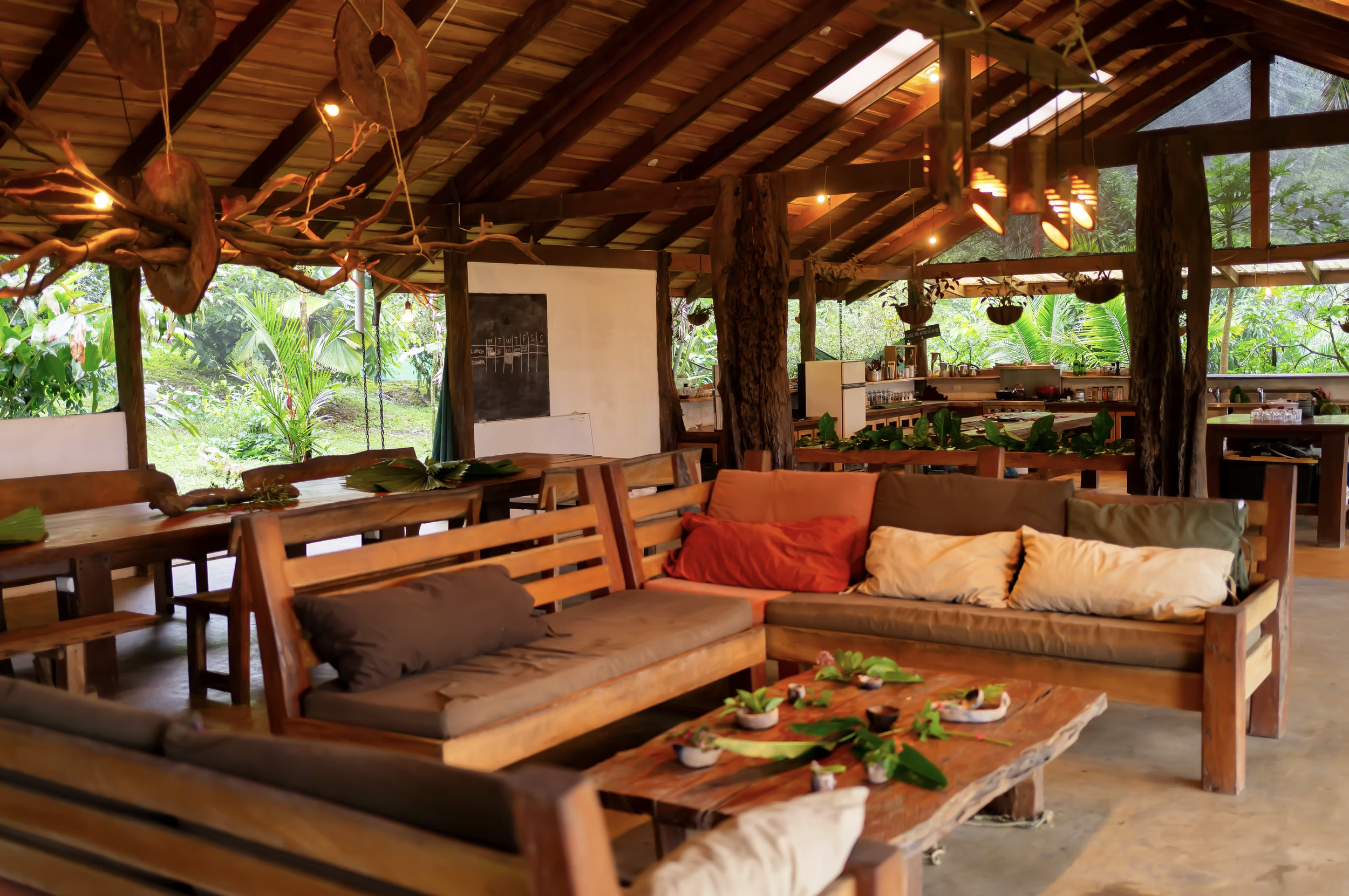 Open-air rustic living space with wooden furniture, a sofa with cushions, and a kitchen area with greenery visible through large windows.