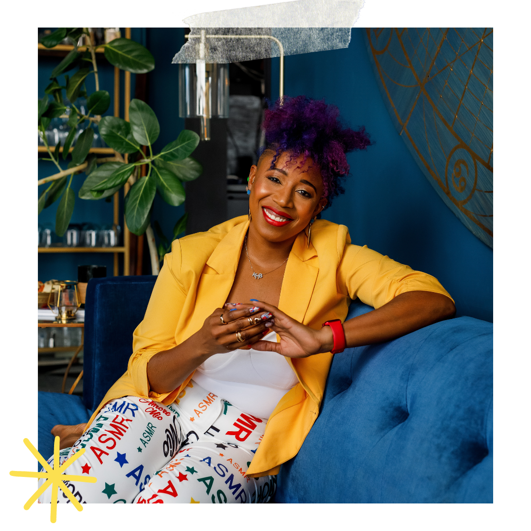 A woman, Dr. Mel Robinson-Brown, with curly purple hair smiling while sitting on a blue velvet couch in a decorated room with plants and art on the walls. She is wearing a yellow blazer, colorful ASMR-themed pants, and jewelry.