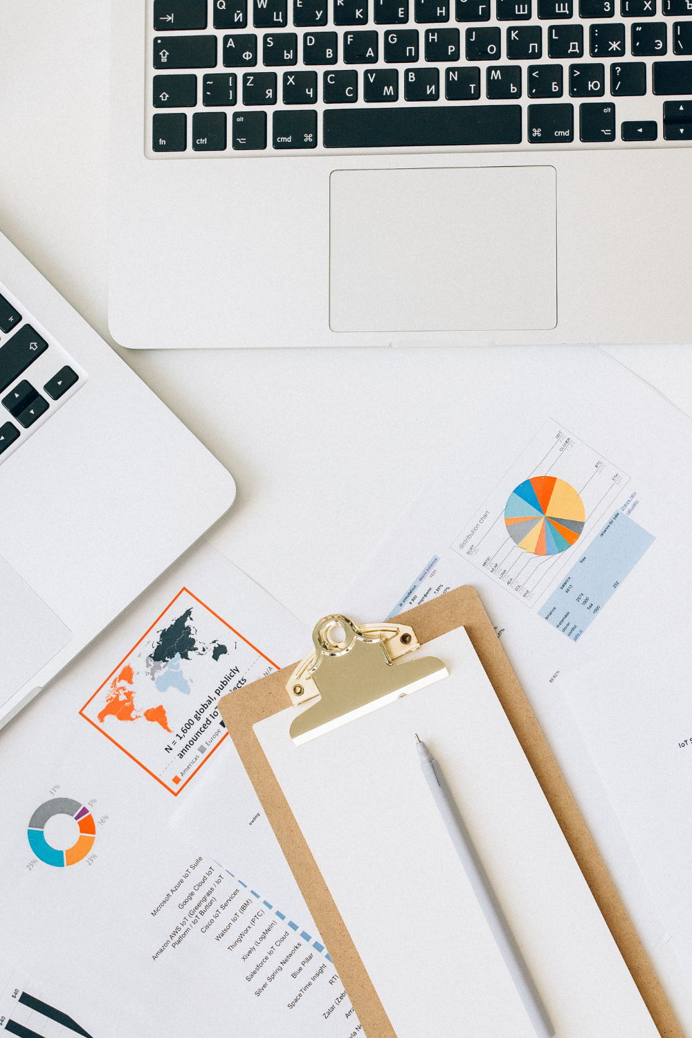 Work desk with two laptops, printouts of charts and maps, a blank notepad on a clipboard, and a pen.