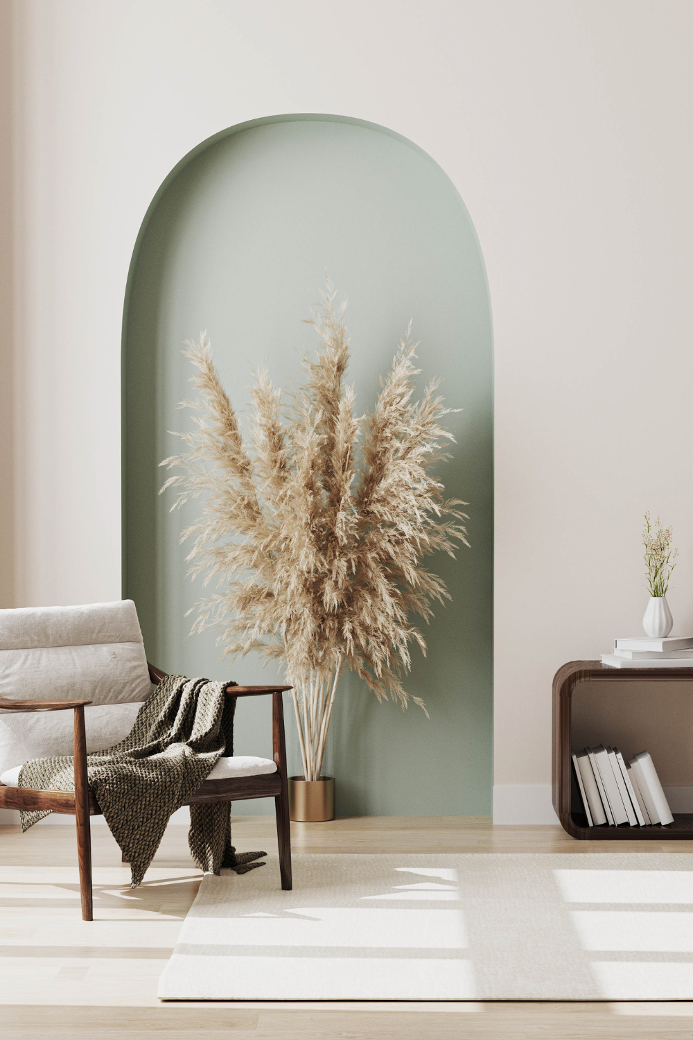 A living room corner with a beige sofa, a wooden armchair draped with a patterned blanket, a large pampas grass arrangement in a gold vase, a small side table with a white vase and books, and a light-colored rug, with a green arched wall niche behind the pampas grass.