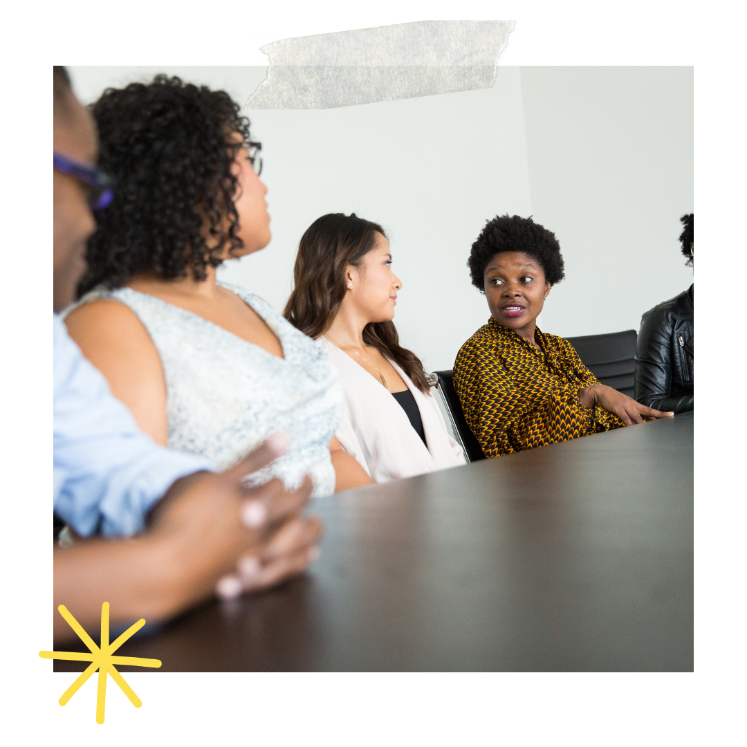 Group of women in a meeting or discussion, sitting at a conference table, one woman speaking.