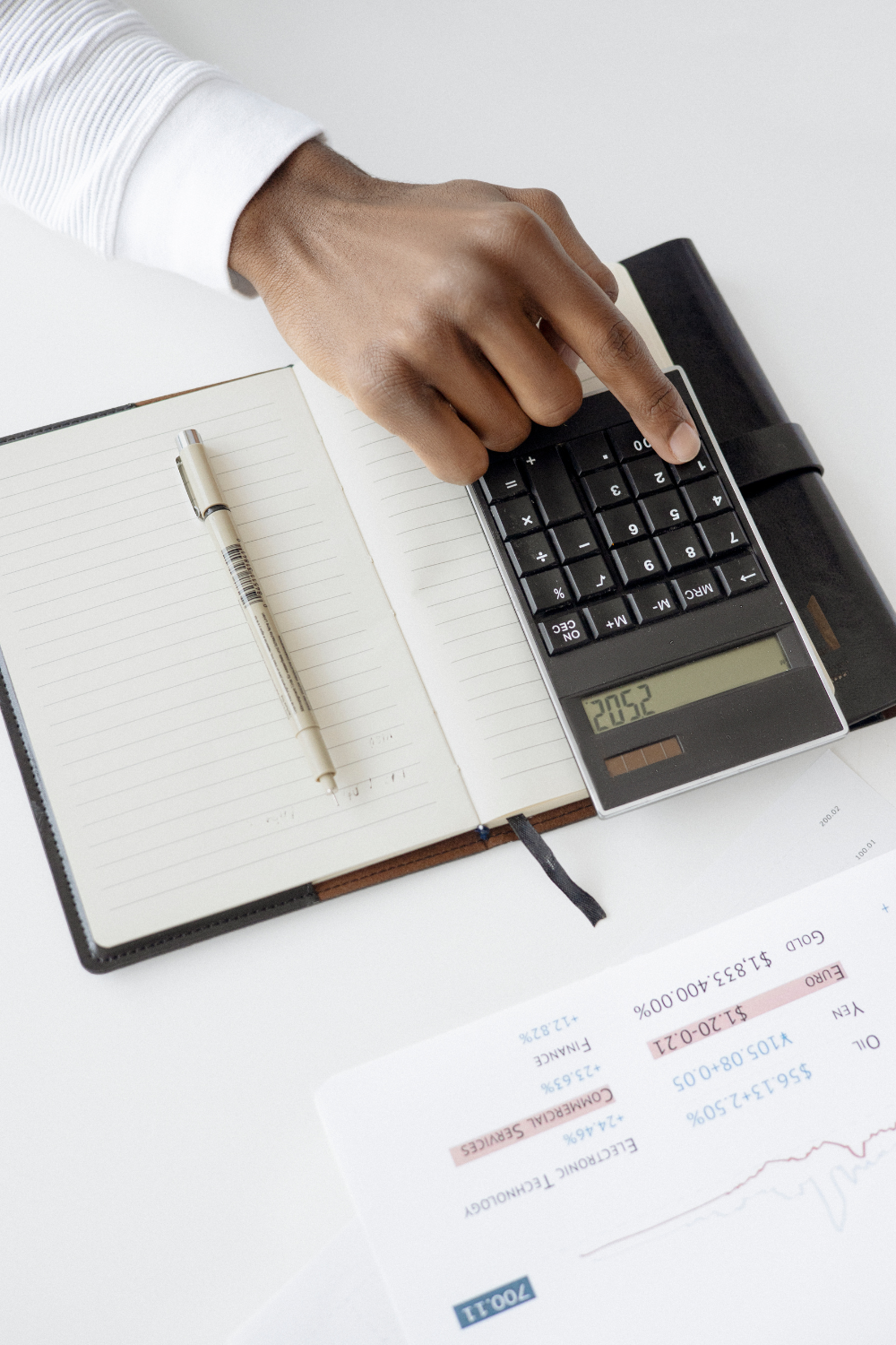 A person's hand using a calculator on a white desk with a notebook and pen nearby, financial documents are also visible.
