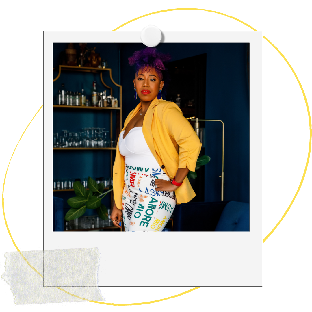 Woman with purple hair in a curly updo, wearing a yellow blazer over a white top and white pants with colorful words, standing indoors with a dark blue wall, shelves with glassware and bottles, and a plant in the background.