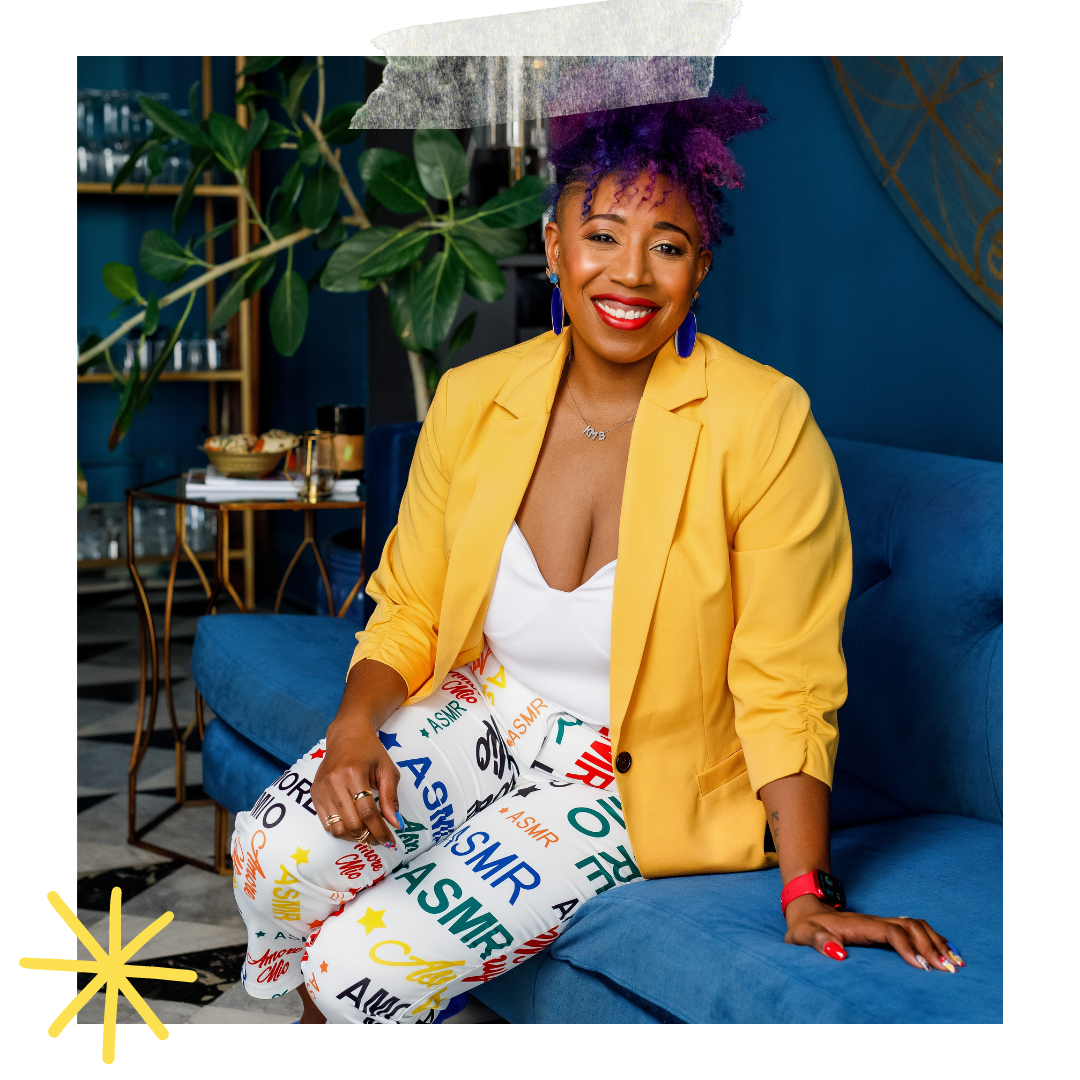 A woman with purple curly hair and a wide smile, sitting on a blue sofa in a stylish room. She is wearing a yellow blazer, white top, and pants with colorful ASMIR and ASMR text. There is a large plant and a shelf with glasses behind her.