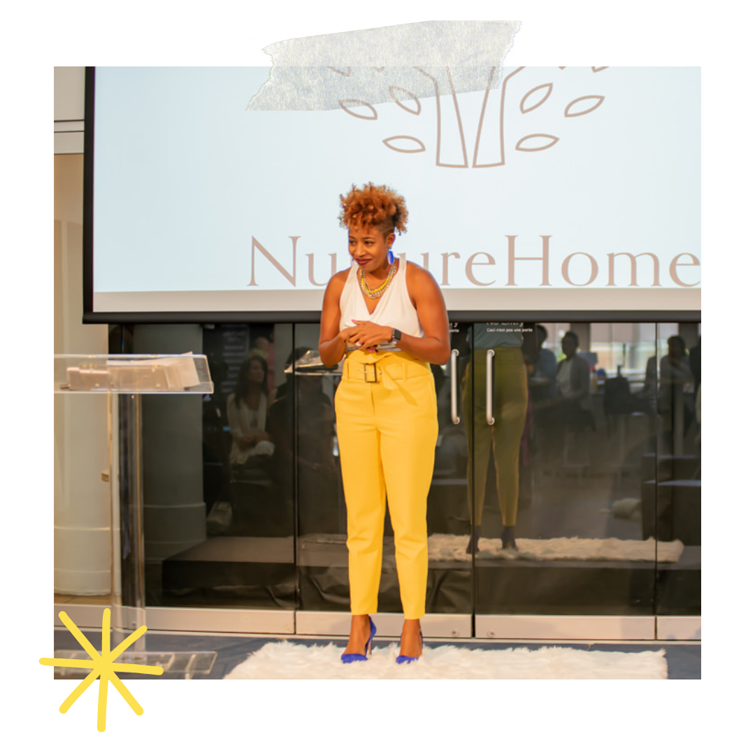 A woman standing in front of a large screen with the logo 'NutureHome' and a tree illustration. She is wearing a white top, yellow pants, and blue high heels, and has short, curly hair.