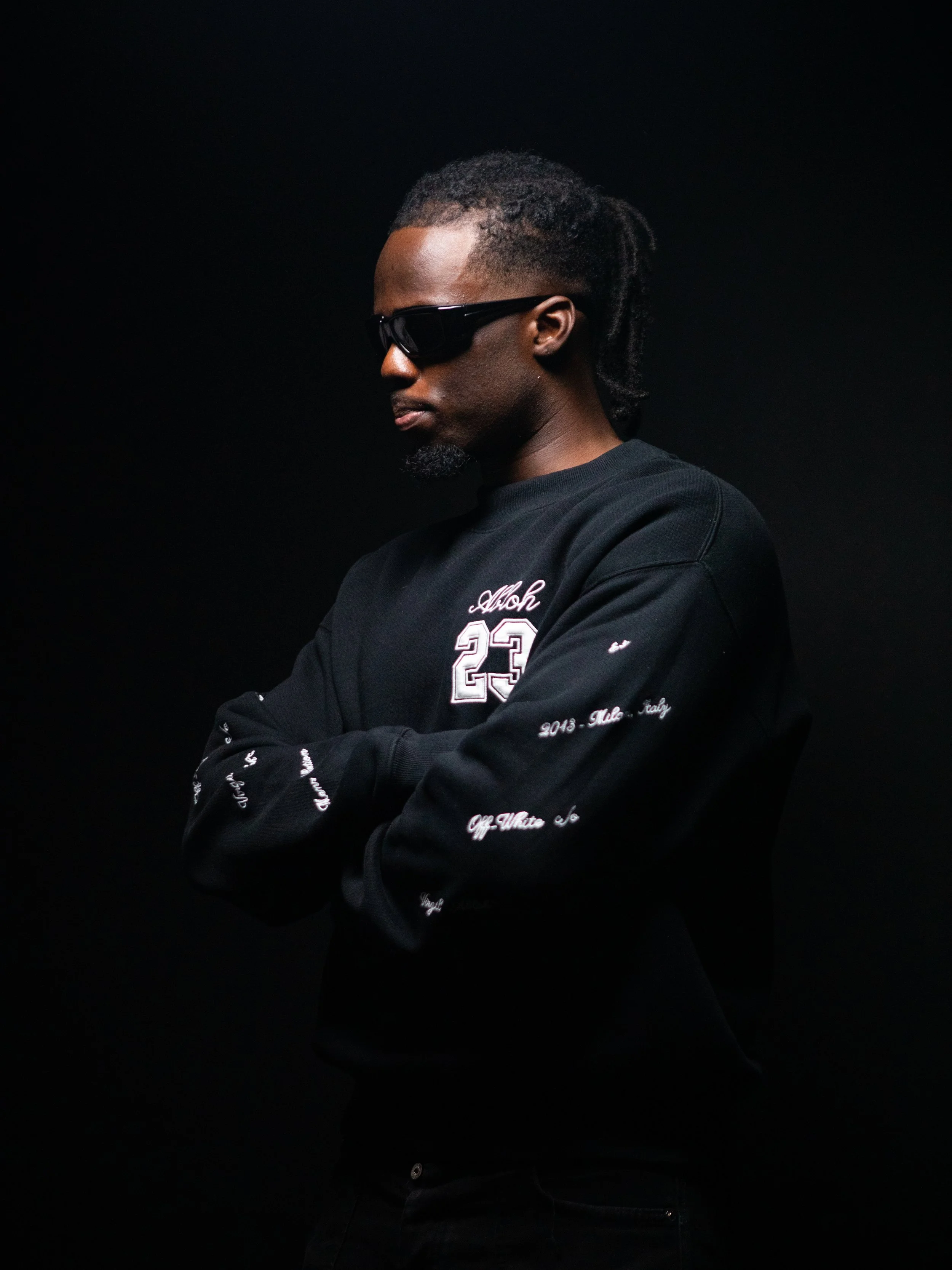 A young man with dreadlocks wearing sunglasses and a black sweatshirt with white embroidery, standing with arms crossed against a dark background. Campaign, Album cover shoot for artist.