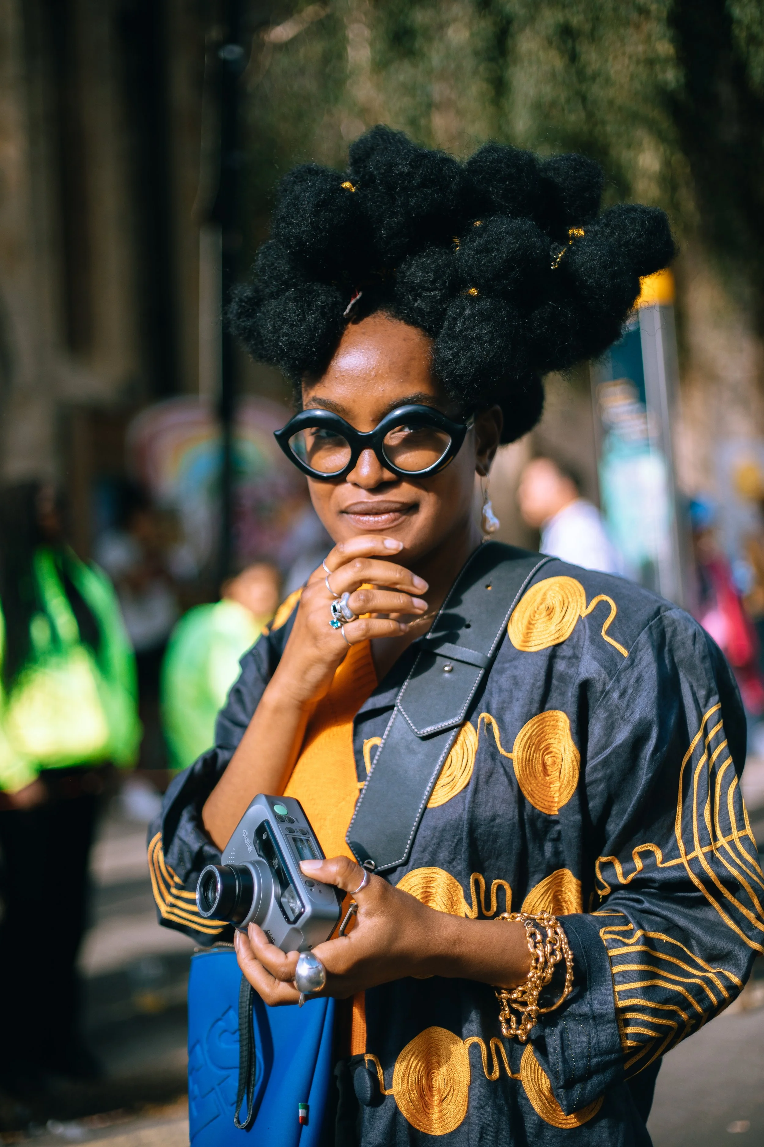 A woman with an elaborate hairstyle and large glasses, holding a camera, standing outdoors during the daytime with a blurred background of people. Notting Hill Carnival int he sunshine.