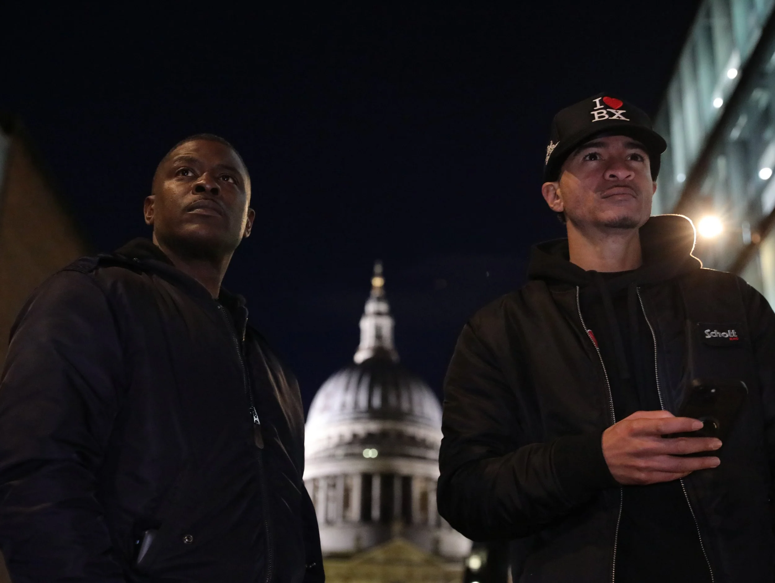 Two men standing outdoors at night, with a domed building in the background, illuminated by lights. One man is holding a phone, both are wearing dark jackets and caps.