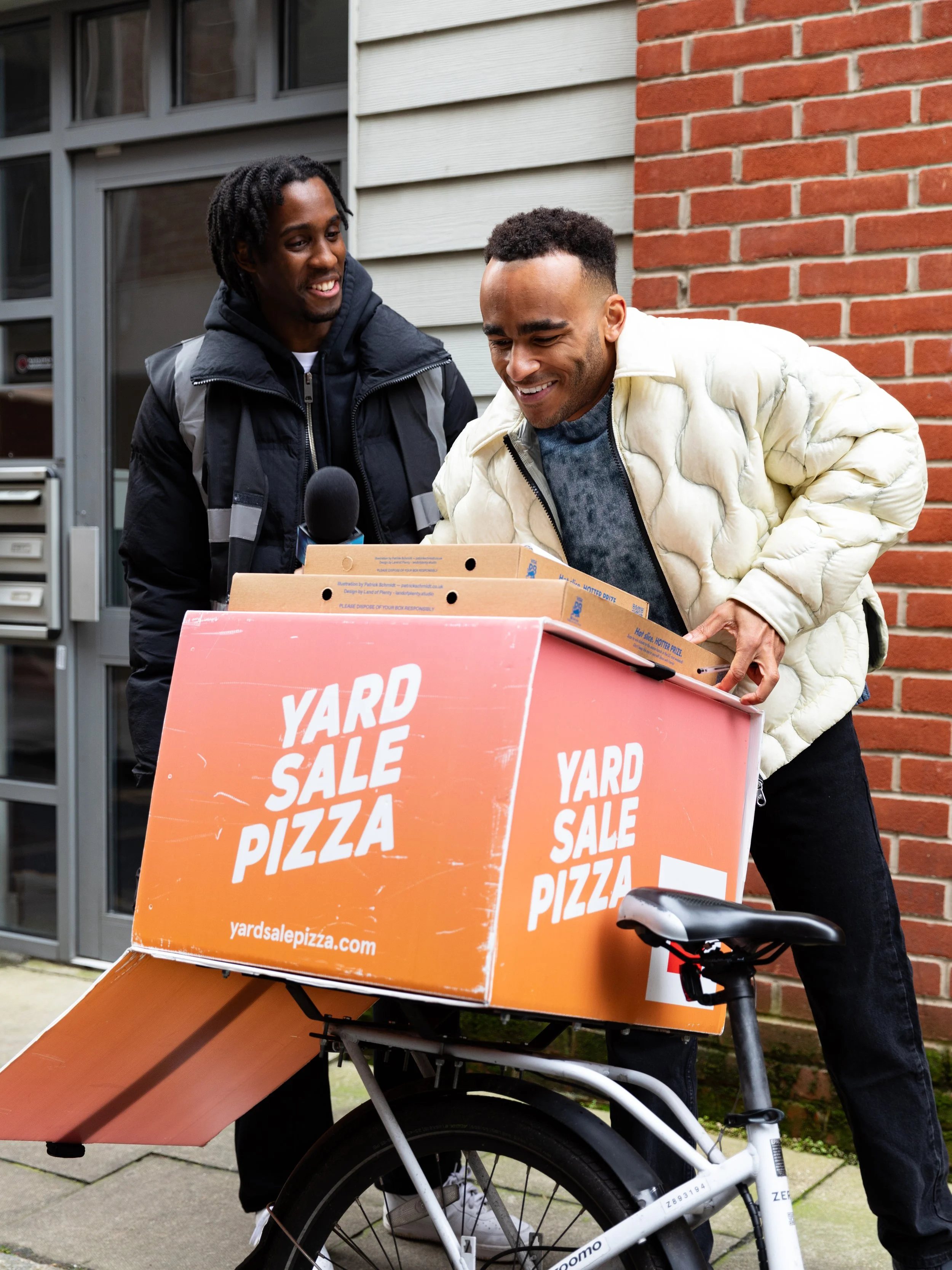 Two men smiling and exchanging items near a bike with a large pink box labeled 'Yard Sale Pizza' on it, outside a building. Behind the scenes for Visa Cash App RedBull, VCARB Social first ad Campaign 