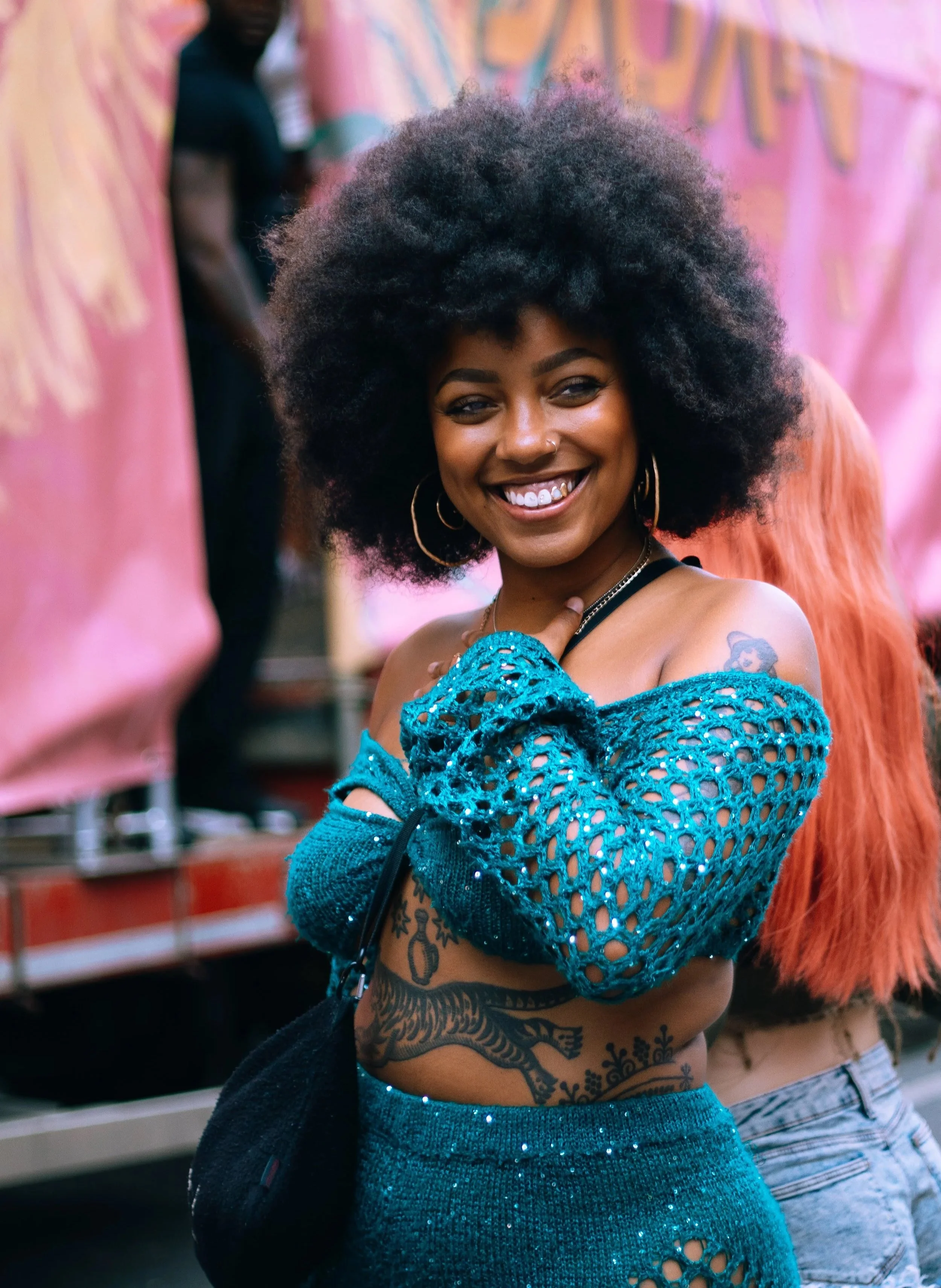 A smiling woman with dark skin, curly afro hair, wearing large hoop earrings and a nose ring. She is dressed in a teal, off-shoulder, crochet top with cutouts and matching high-waisted shorts. She has tattoos on her stomach and arm, and is standing i