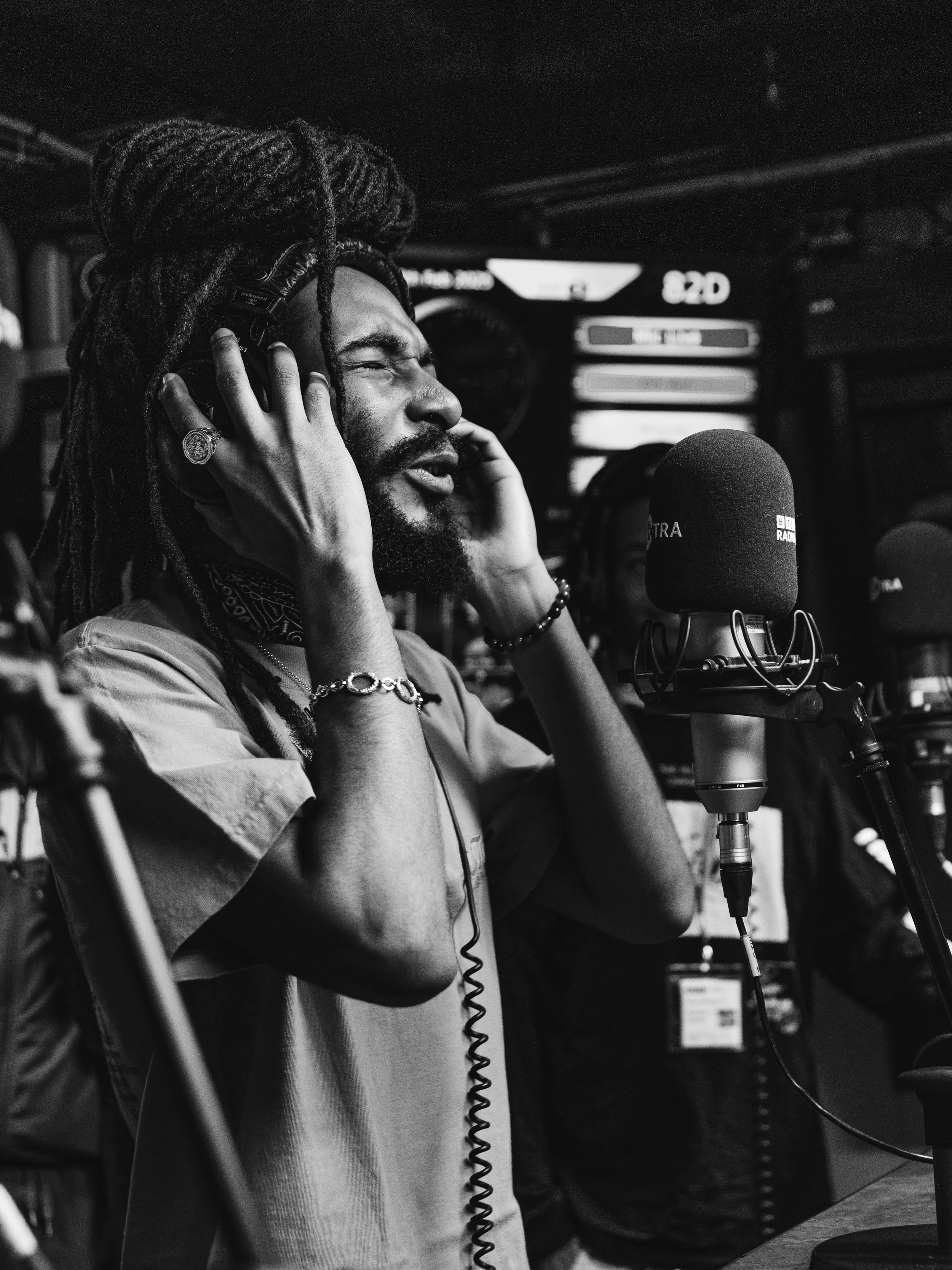 A man with dreadlocks and a beard in a recording studio, wearing headphones, singing or speaking into a microphone.