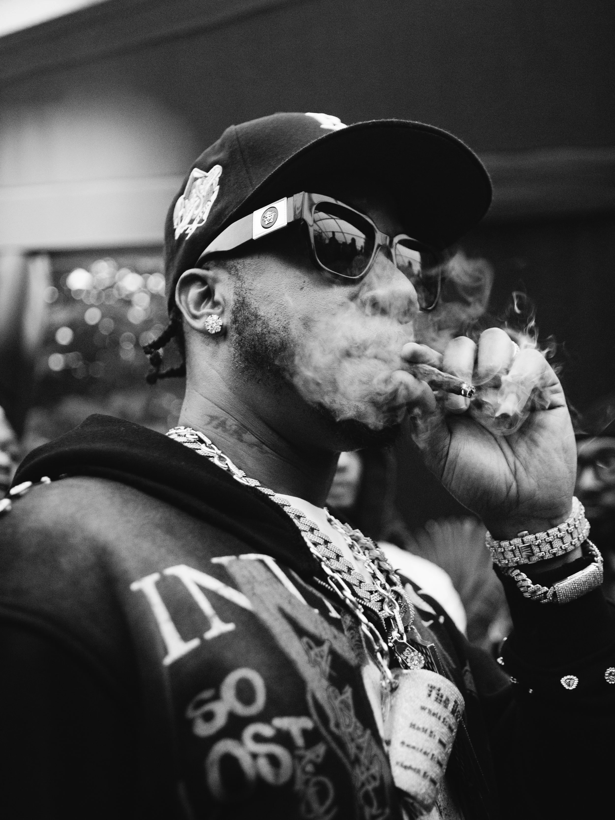 A man, Benny the Butcher, wearing a baseball cap, sunglasses, and jewellery, smoking a Blunt in a social setting. Backstage and Jazz Cafe Festival, London.