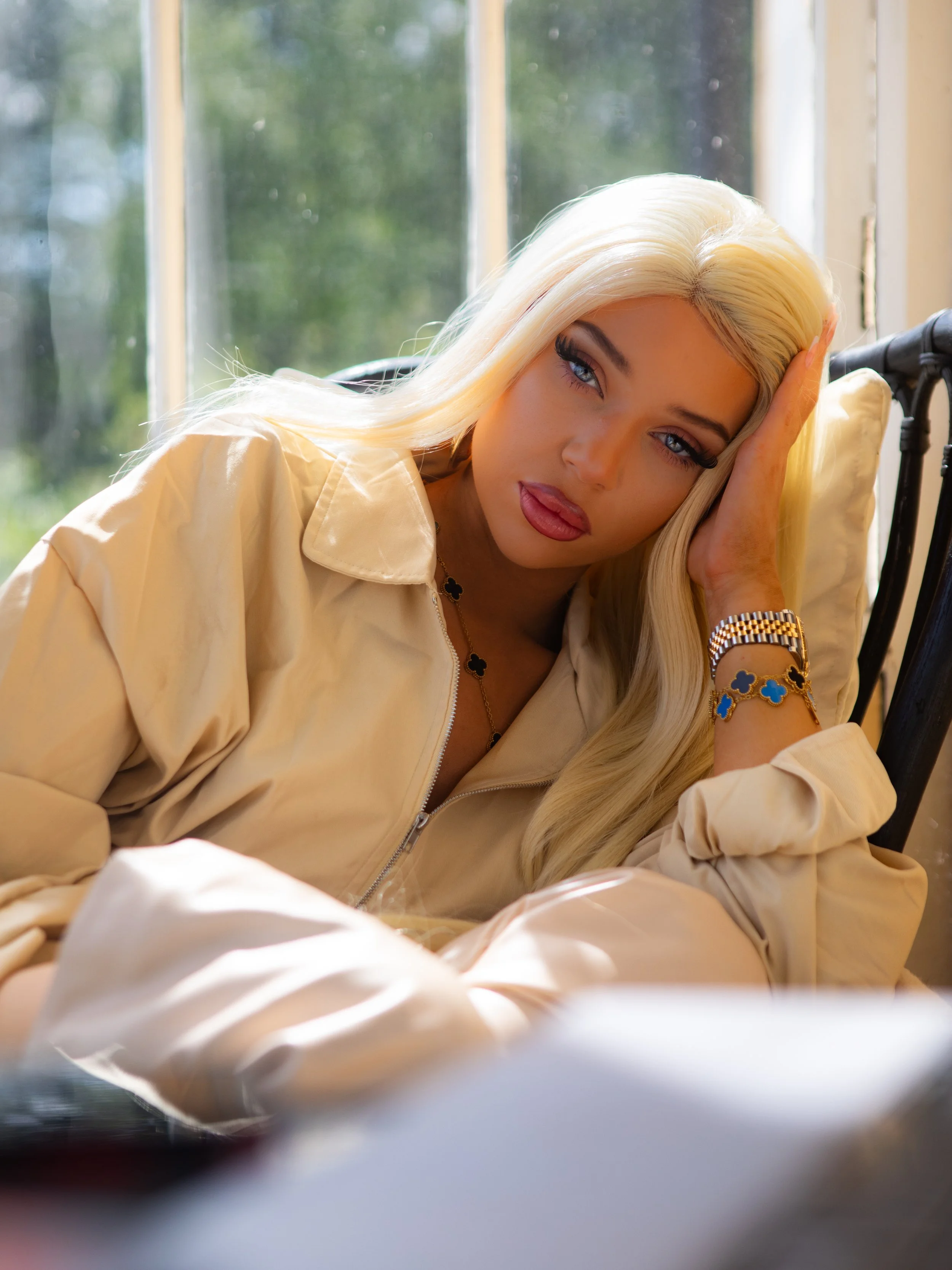 A woman with long platinum blonde hair and blue eyes resting her head on her hand while sitting by a window on a sunny day in Stockholm, Sweden.