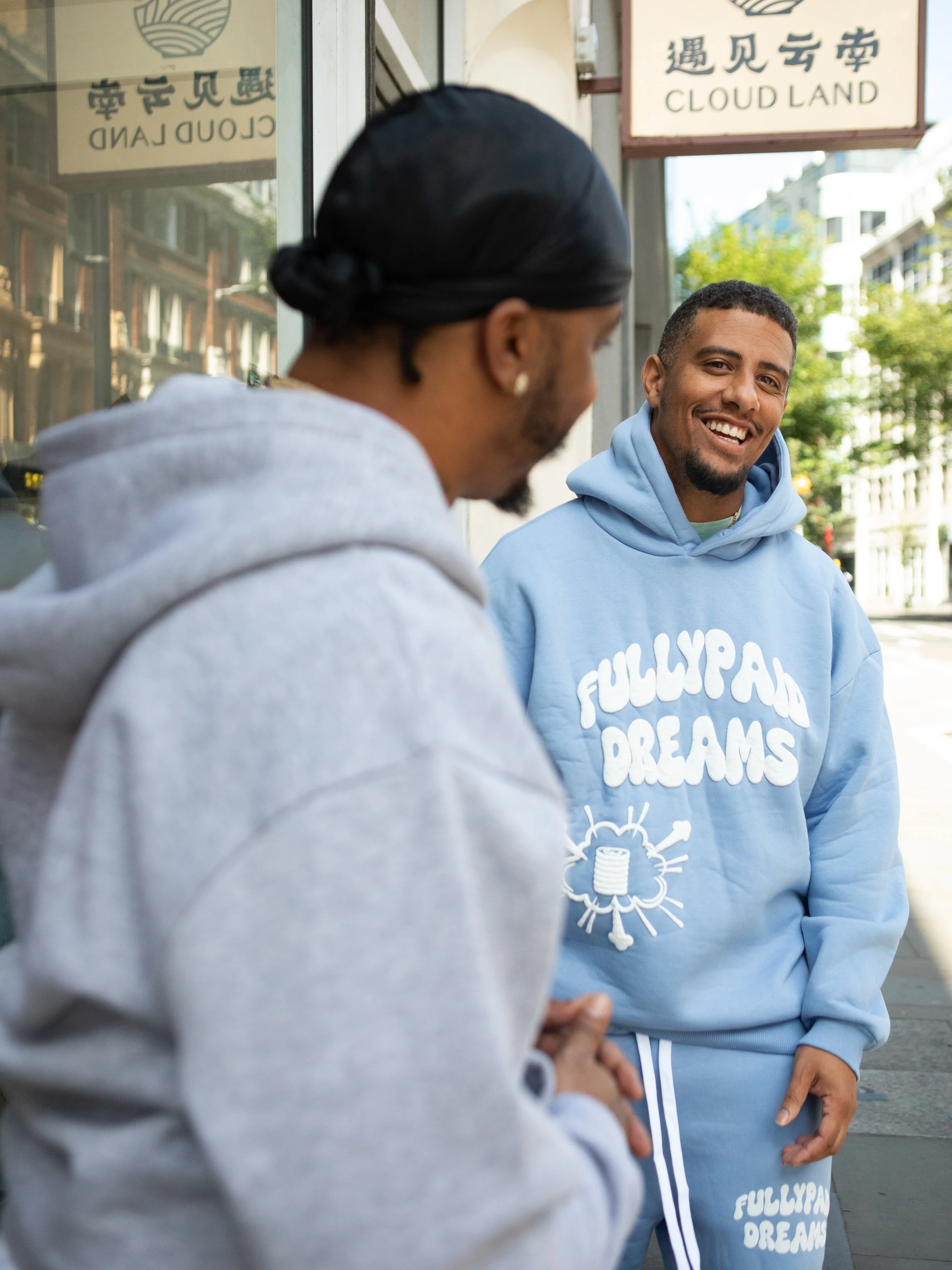 Two men are standing outside on a London Street, one facing away in a gray hoodie, and the other smiling and looking at him in a blue hoodie with 'FULLY PAID DREAMS' printed on it. Benny The Butcher and his manager Jake Amankwaah