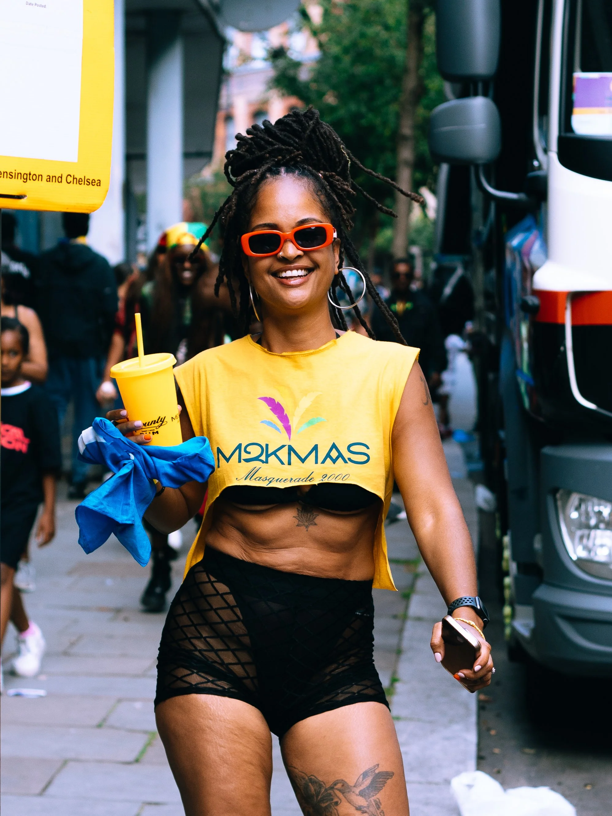 A woman with dreadlocks, red sunglasses, and hoop earrings smiling at a street festival. She is wearing a yellow crop top with a colorful logo and black fishnet shorts. Behind her, there are other people and a black truck and Notting Hill Carnival