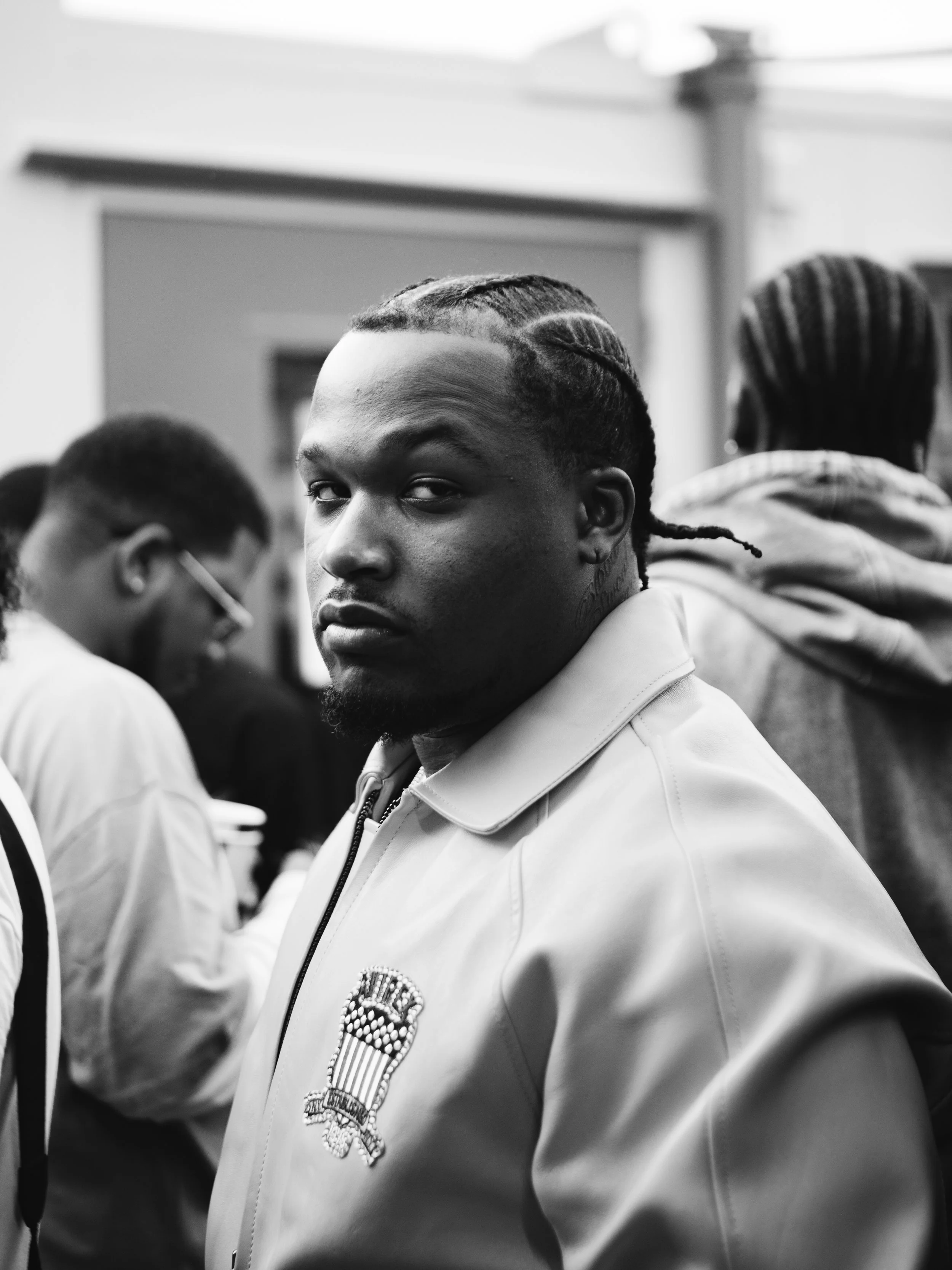 A black and white photo of a young man with braided hair and tattoos, wearing a jacket with an emblem on it, in a crowded indoor setting. ElCamino, B$F, Backstage at Jazz Cafe Festival, London, In a Custom Avirex Jacket.