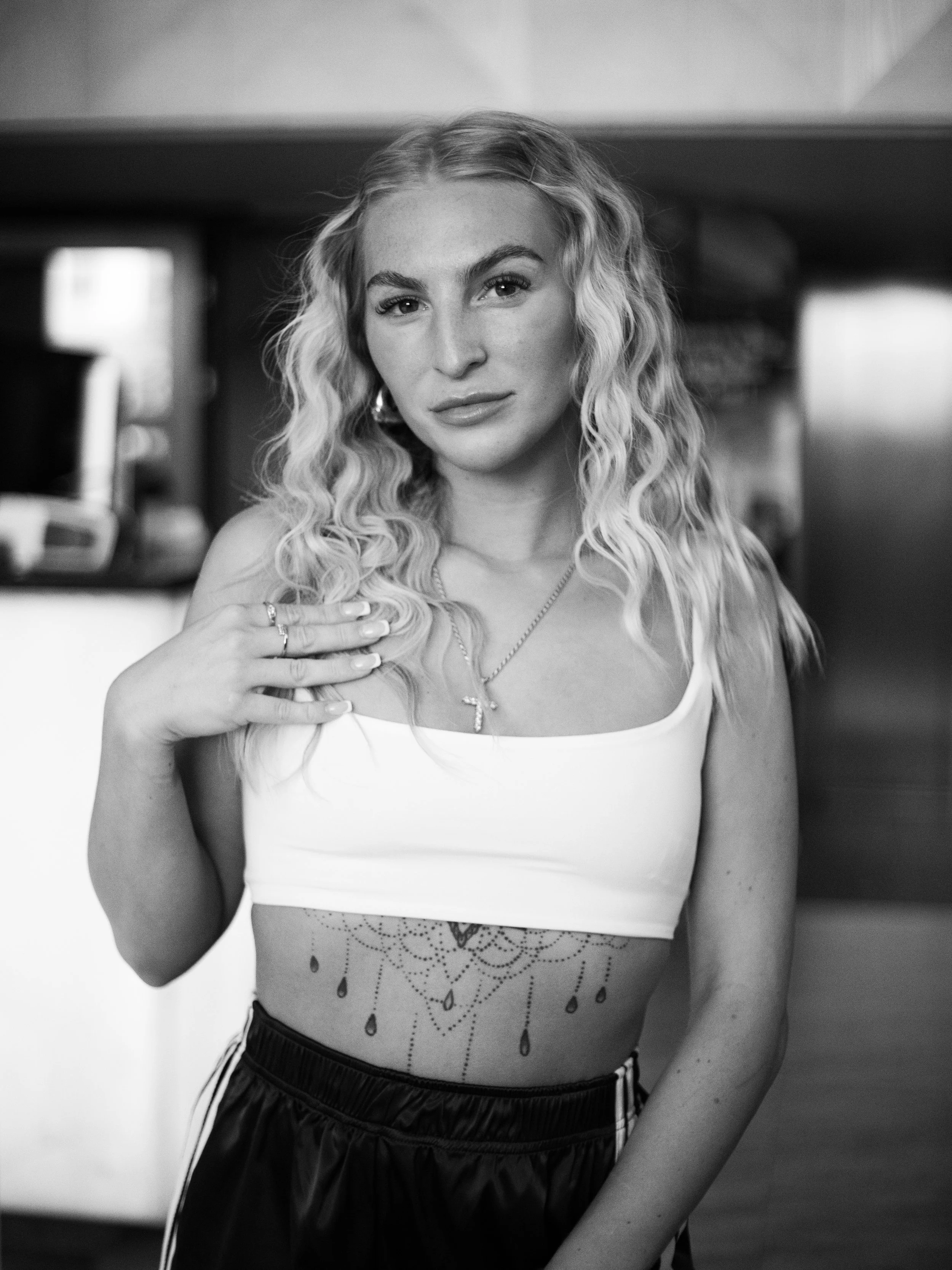 A woman with long, curly blonde hair, wearing a white crop top and black athletic shorts, standing indoors with a tattoo on her midriff. She has her left hand on her shoulder and is looking at the camera.