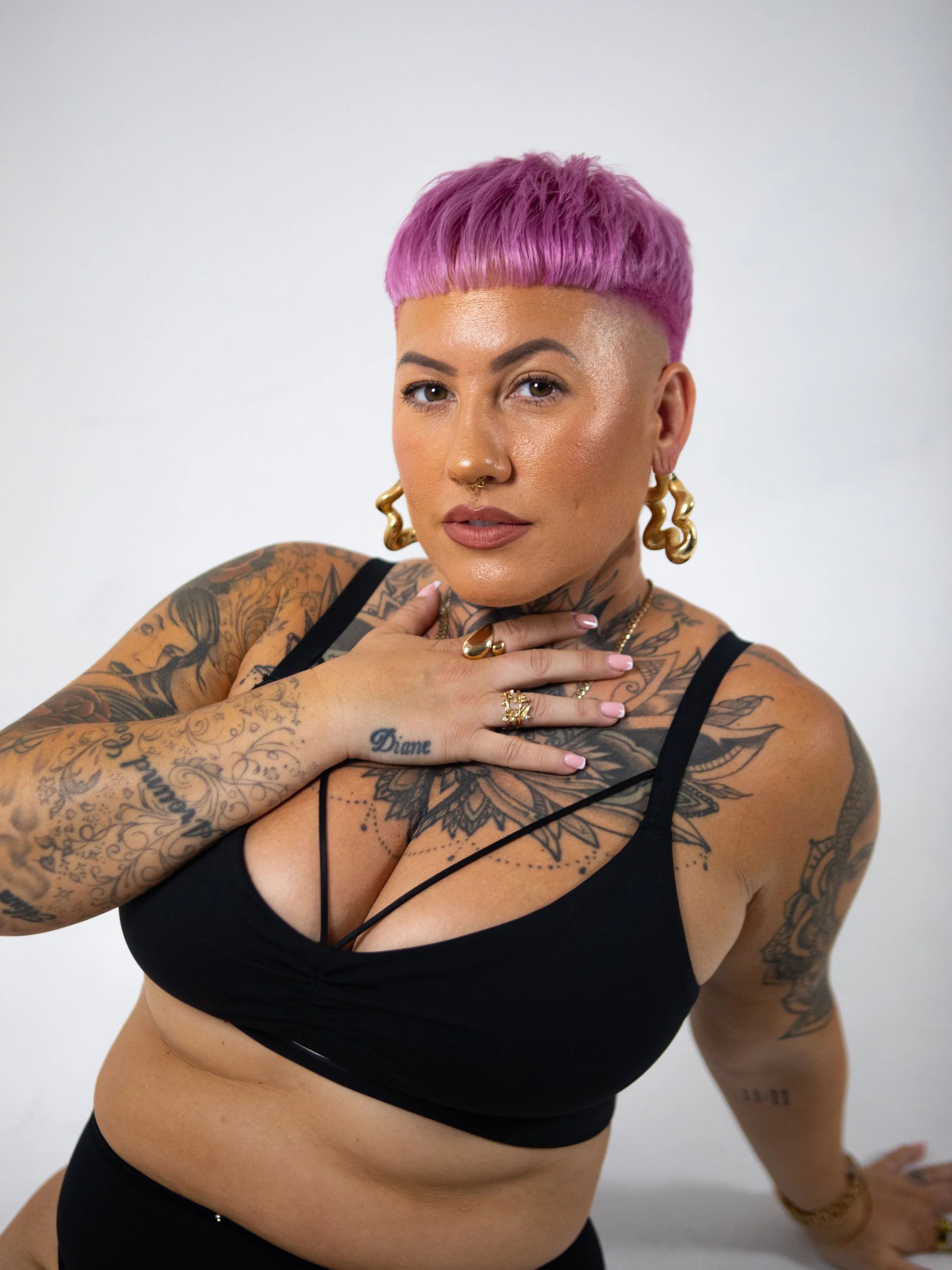 A woman with pink hair, tattoos, and jewelry poses with her hand on her chest against a white background.
