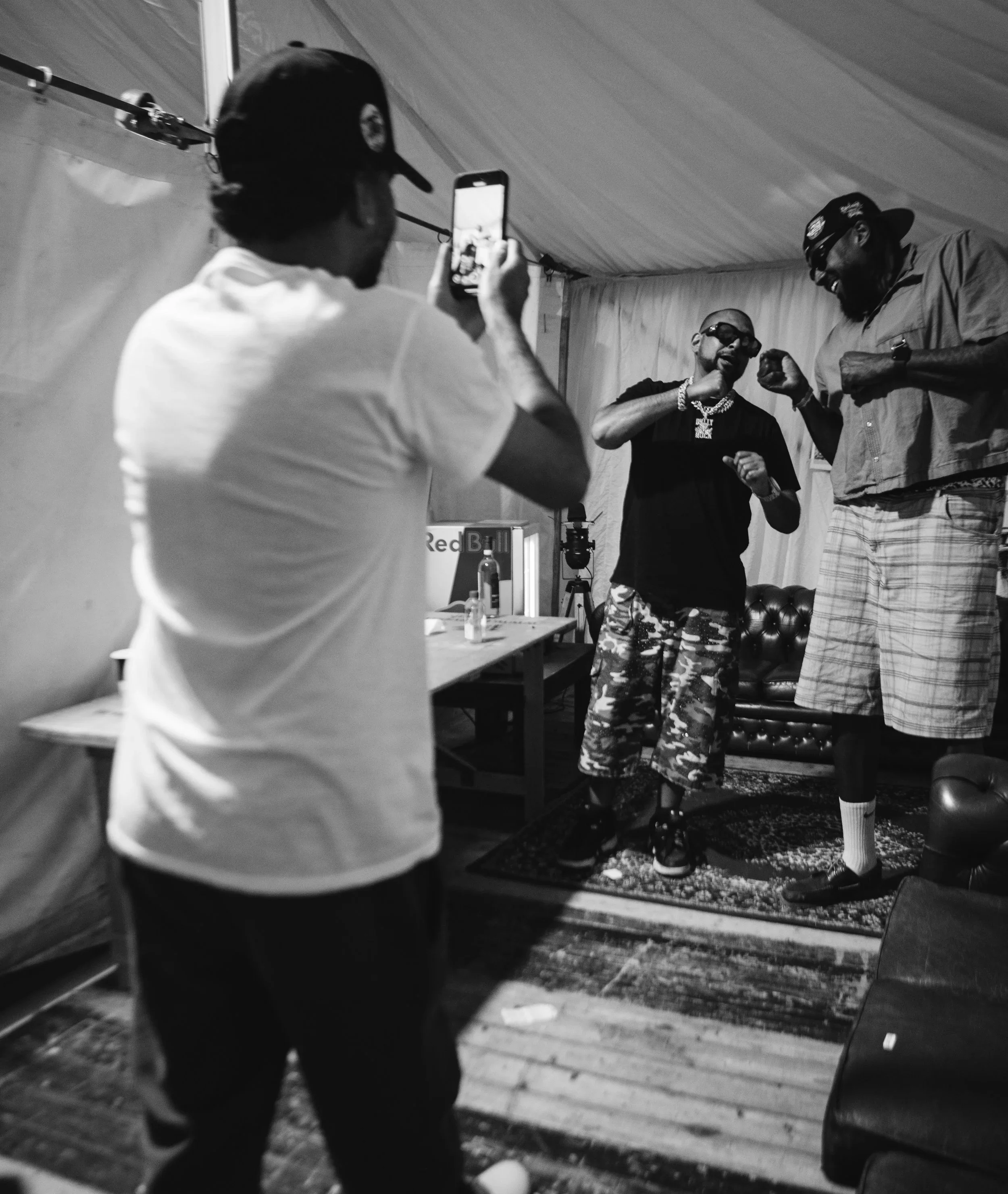 Two men are posing for photos while a person in the foreground captures their image with a smartphone. The men are indoors, smiling, and appear to be in a casual setting with furniture and a carpeted floor.