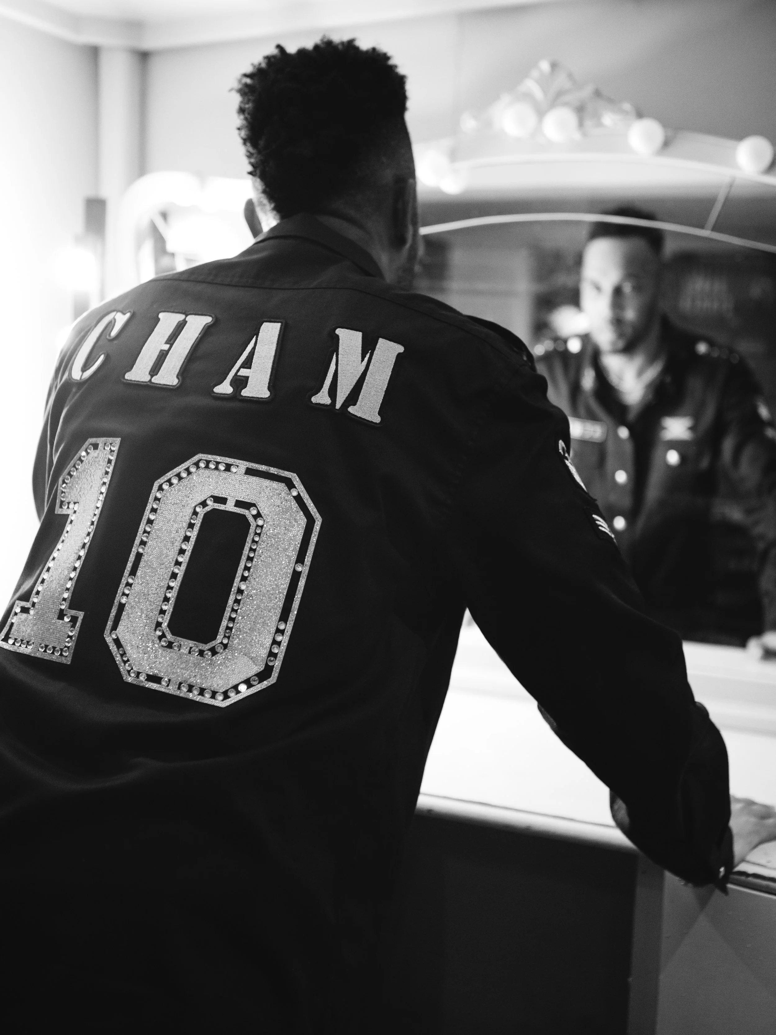 Back of a man wearing a varsity jacket with the name 'Chalm' and the number '10' in rhinestones, looking into a mirror with a woman in uniform in the background.