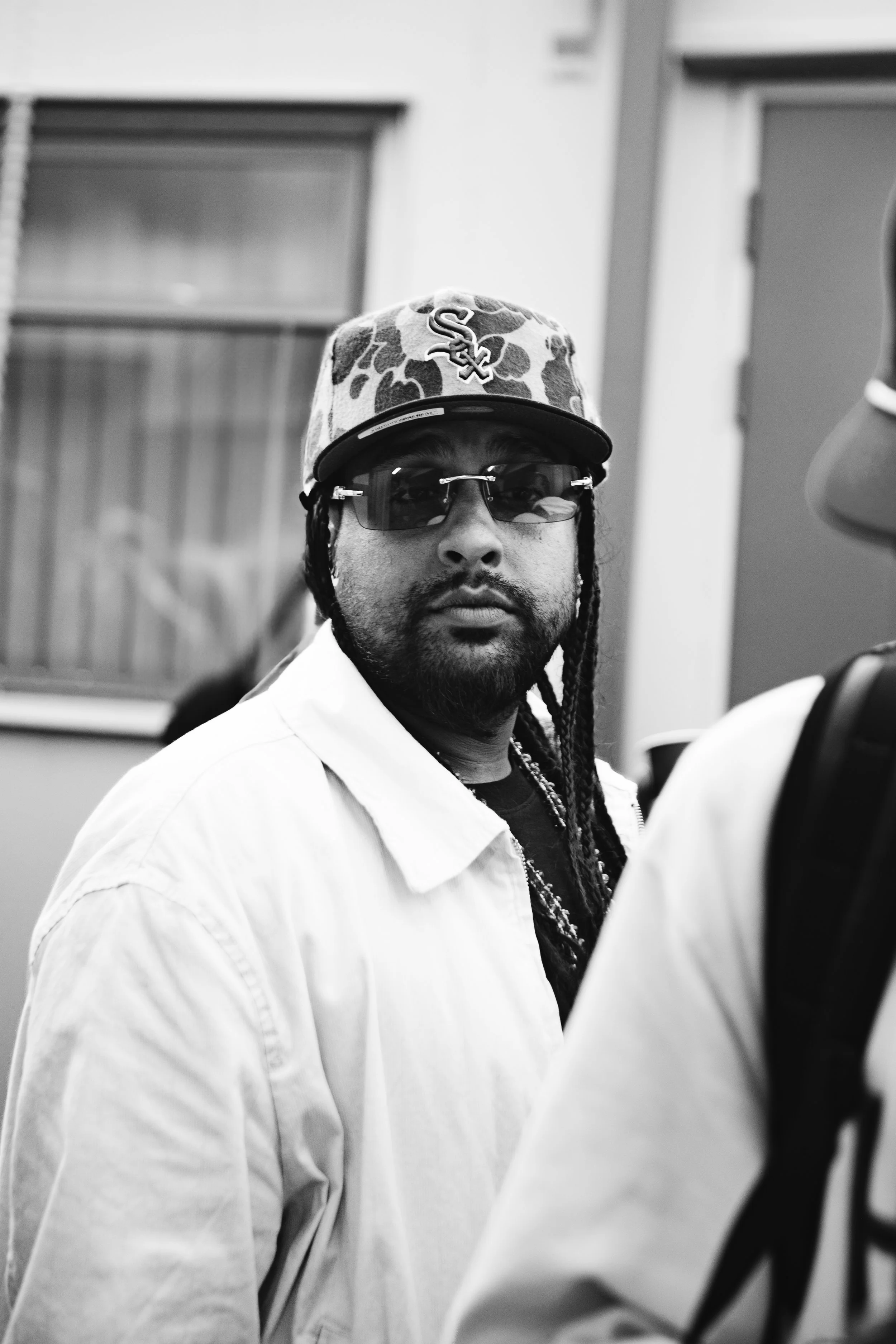 A man in sunglasses and a leopard-print cap with the Chicago White Sox logo, wearing a white shirt, standing outdoors.