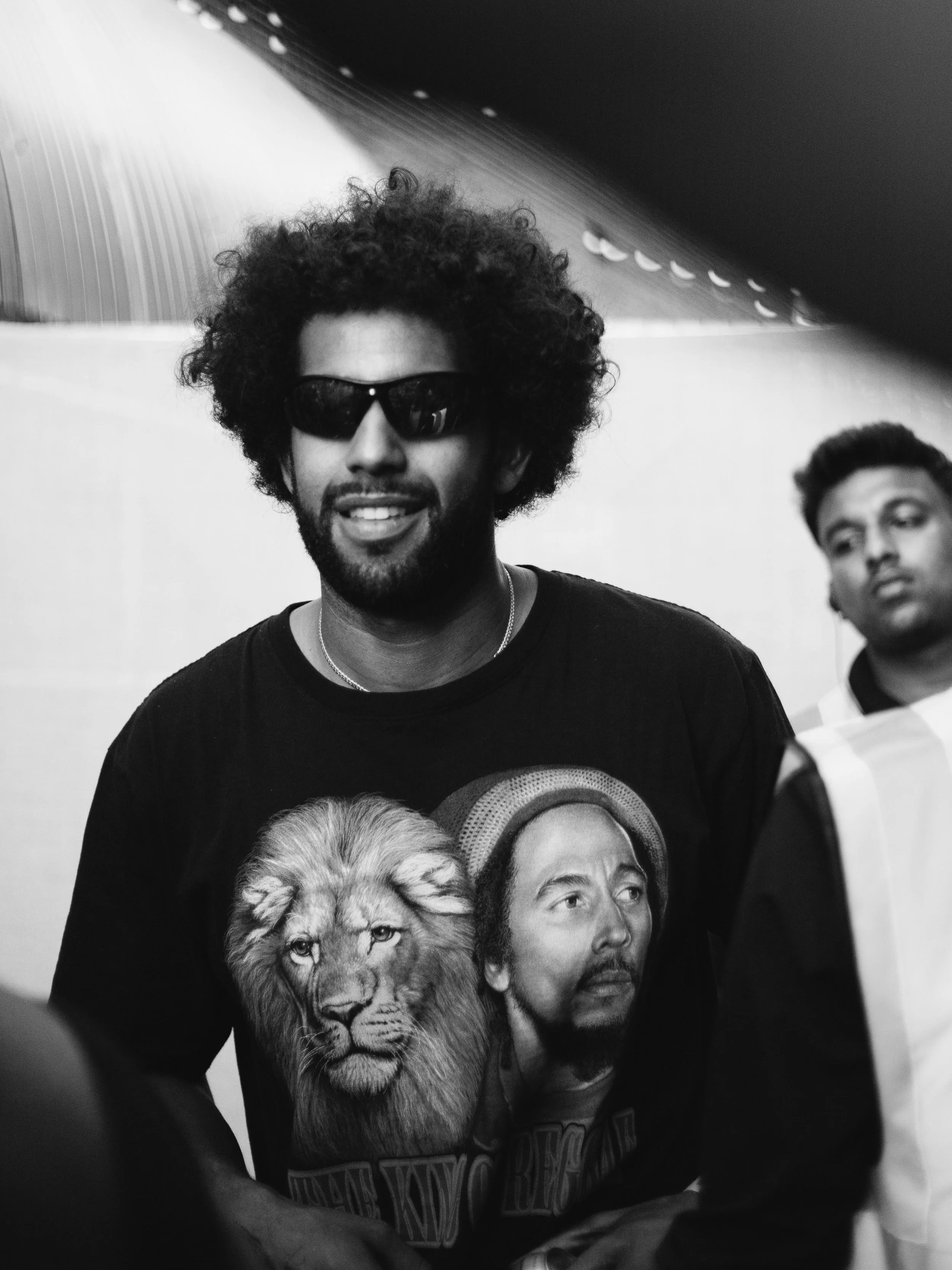 Black and white photo of a man with curly hair and sunglasses smiling, wearing a T-shirt with a lion and Bob Marley on it. Another person is partially visible to the right, and the background features a metallic, ribbed structure.