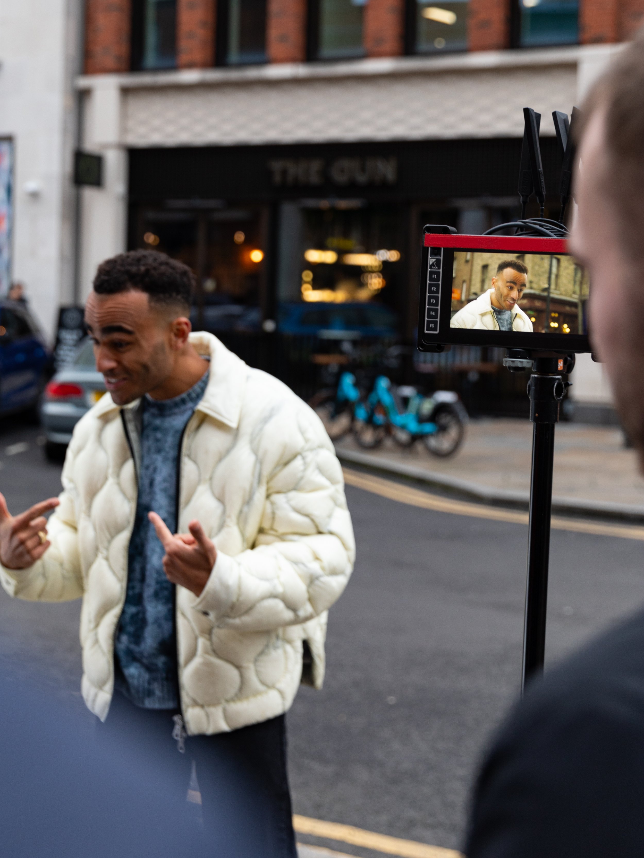 A man in a white quilted jacket is being filmed outside on a city street, seen on a monitor in front of a crew member. The background shows parked cars, motorcycles, and a building with a sign that reads 'The Gun'. BTS for Redbull Campaign.