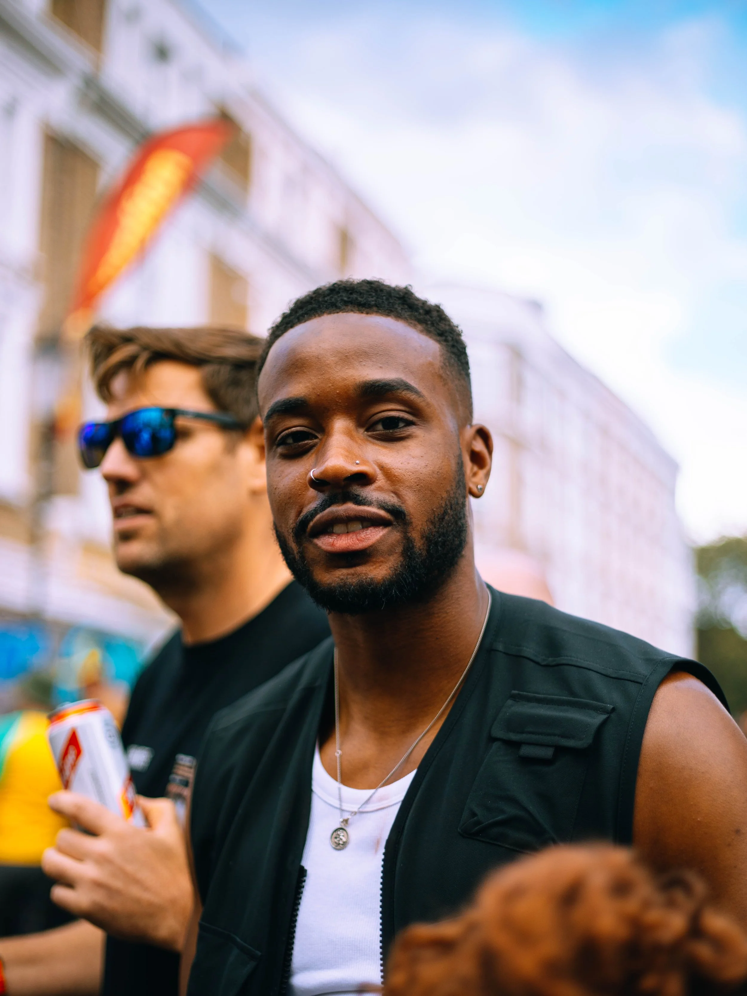 A young man with a beard and earrings looking at the camera, wearing a black sleeveless shirt and a necklace, standing outdoors with an urban background. A second person in sunglasses holding a soda can is slightly out of focus behind him.