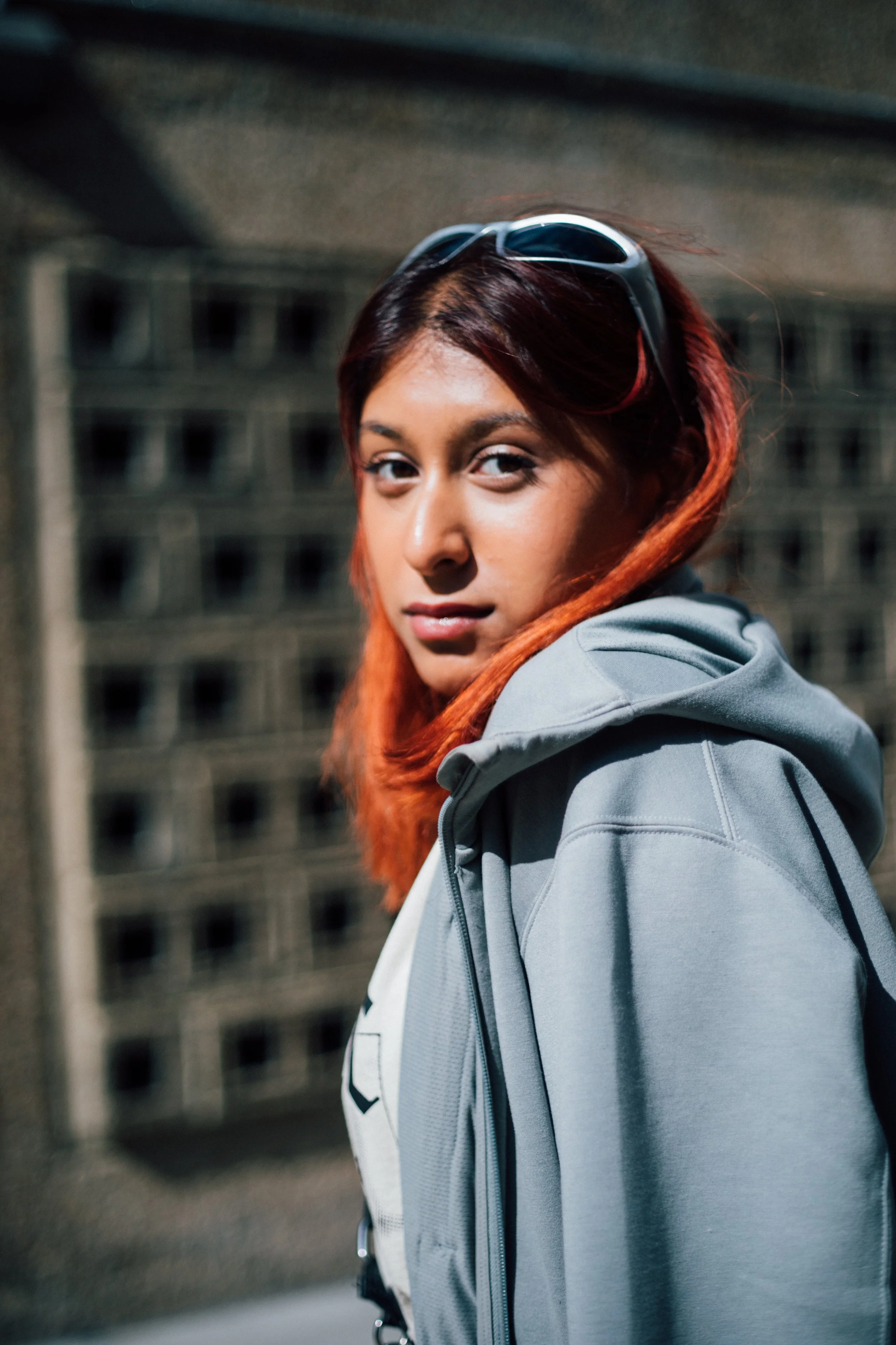 A young woman with reddish-brown hair, wearing sunglasses on her head and a gray jacket, looking over her shoulder at the camera with a blurred cityscape background.
