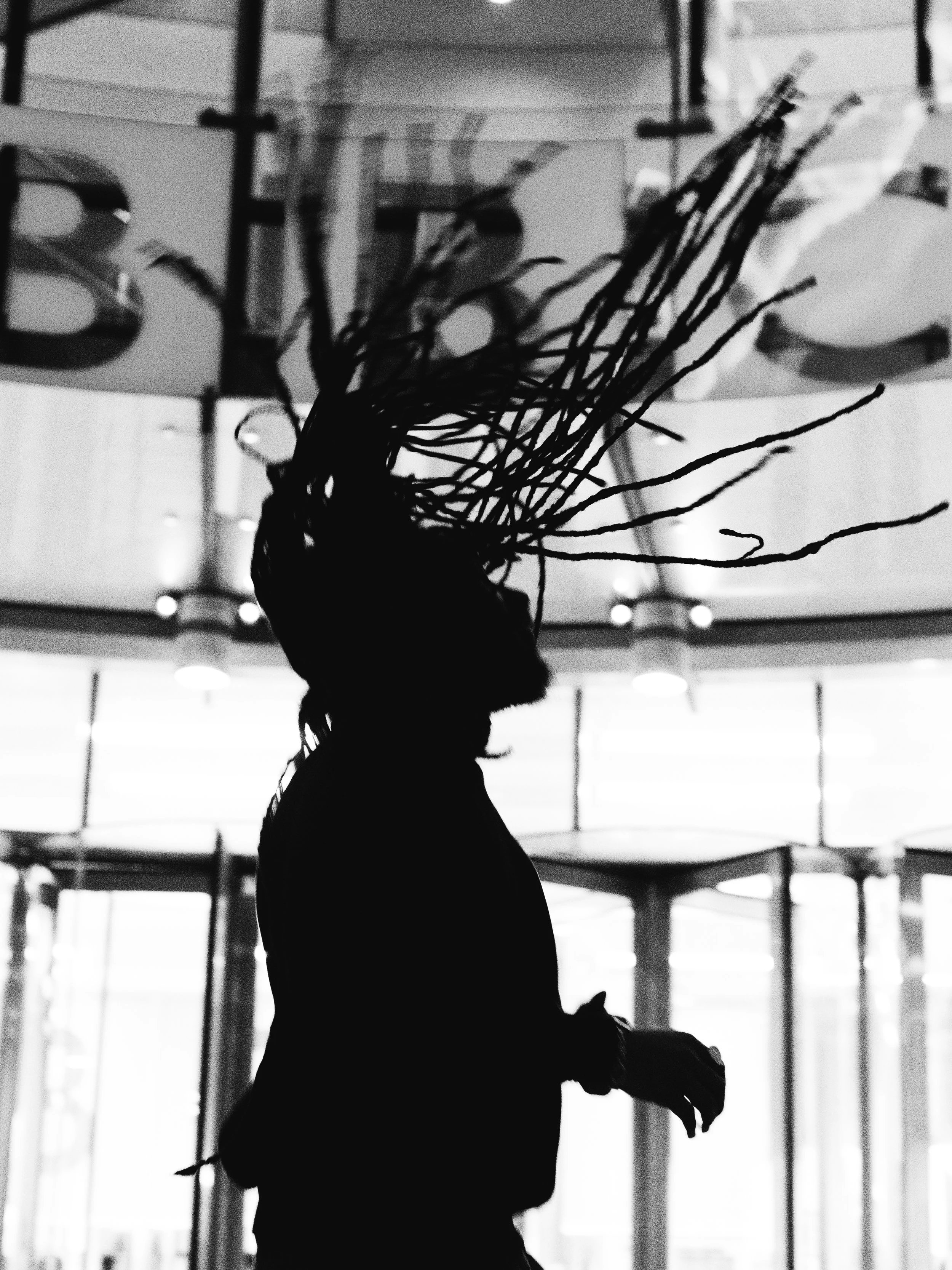 Silhouette of a woman with dreadlocks standing indoors in front of glass doors, with bright light behind her.