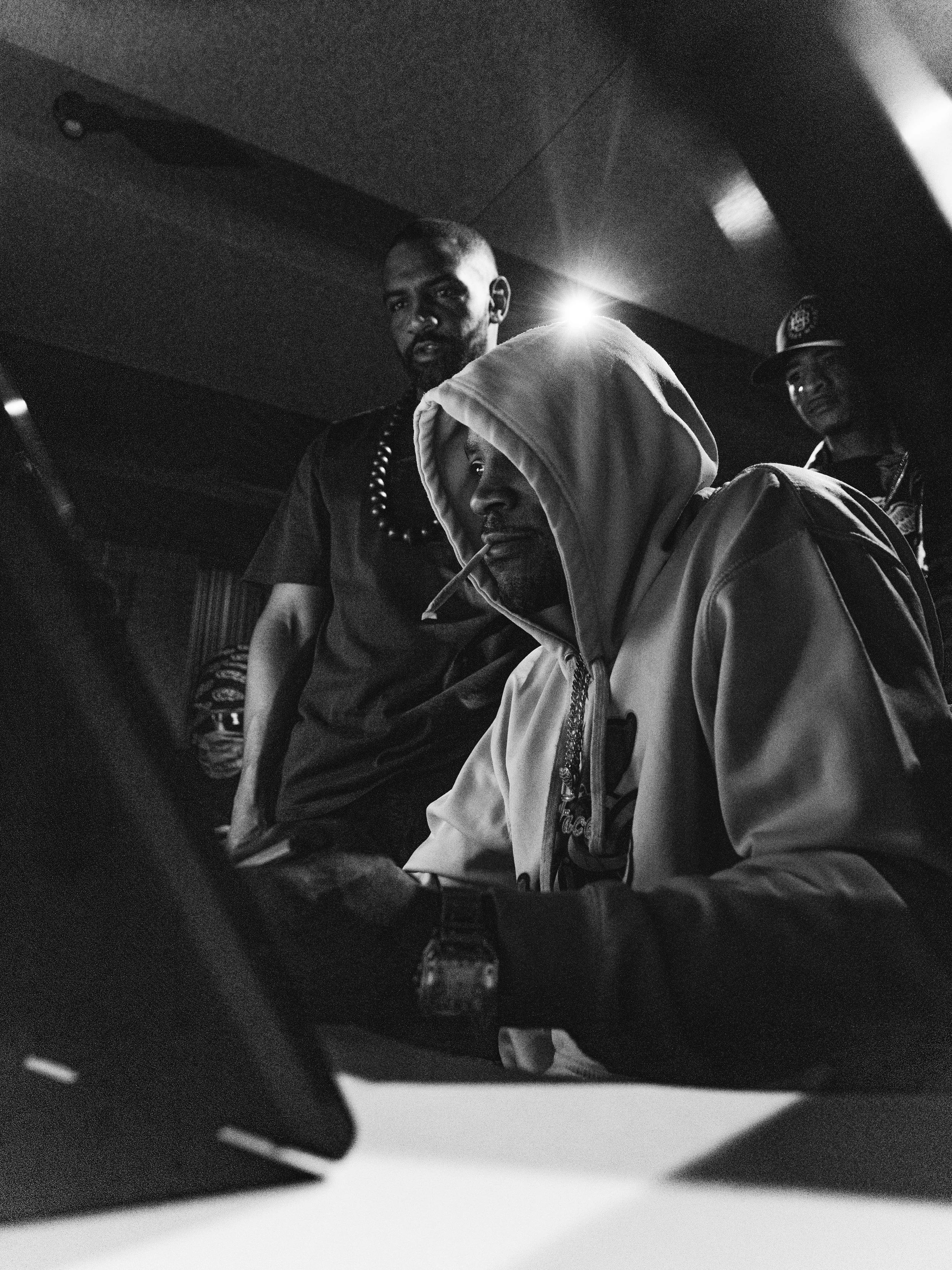 Black and white photo of three men; one sitting with a hood, cigarette in mouth, and looking at a device, two others standing behind him, one with a shaved head and beads, the other wearing a cap, in a dimly lit recording Studio.