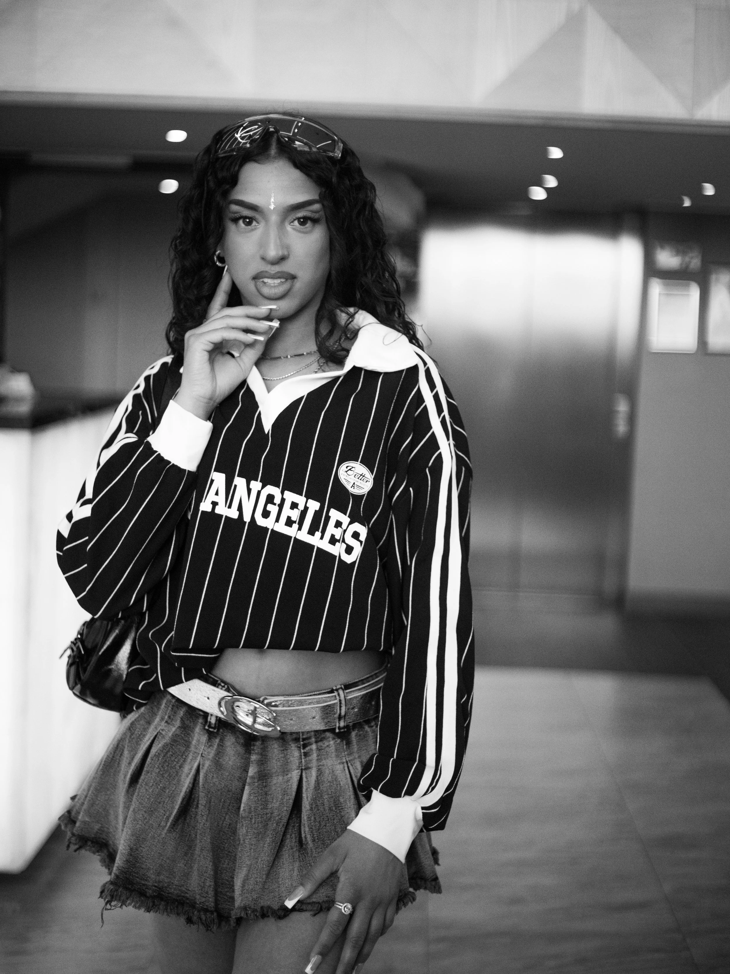 A woman wearing a Los Angeles jersey, pleated shorts, and jewelry stands in a modern indoor setting. She has dark curly hair, a white line on her forehead, and is looking into the camera with her finger on her temple.