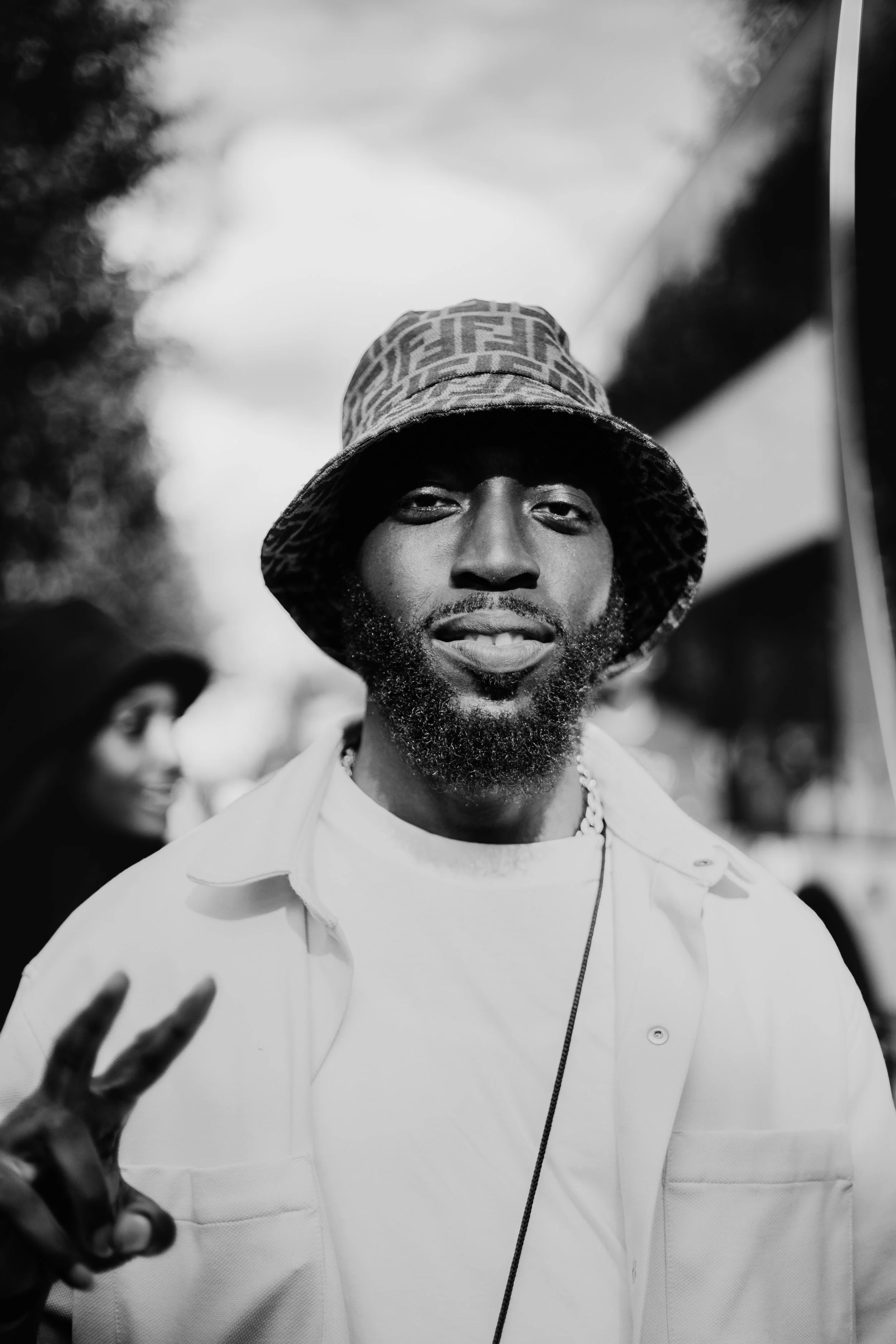 A man with a beard wearing a bucket hat and a light-colored jacket, making a peace sign with his hand.