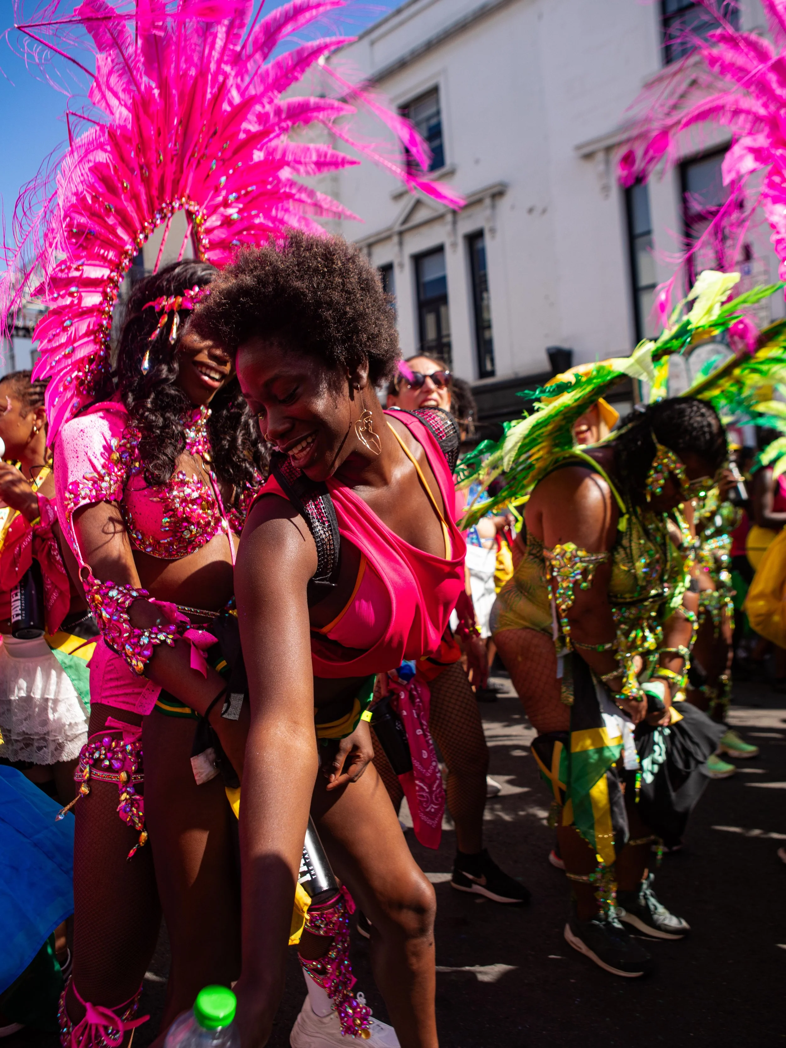 Women dancing in colorful costumes during a parade, Notting Hill Carnival, with pink feathers and sunshine.